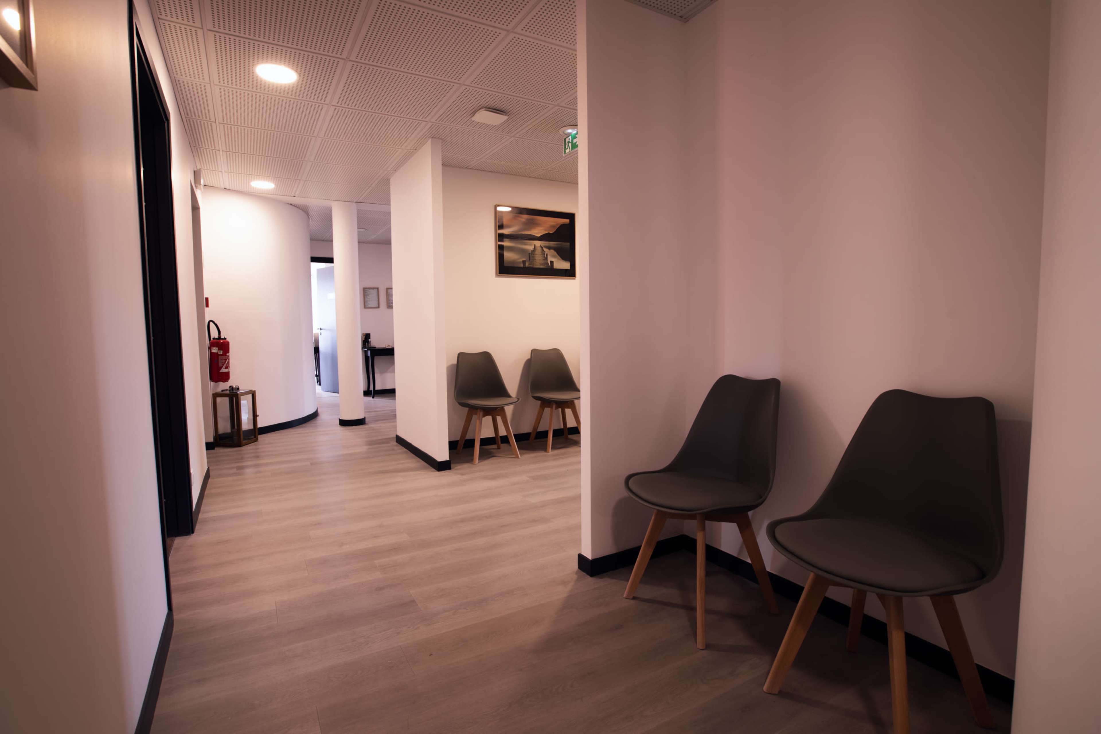 A hallway features light-colored flooring and walls, with chairs along one side and an open area leading to another room.