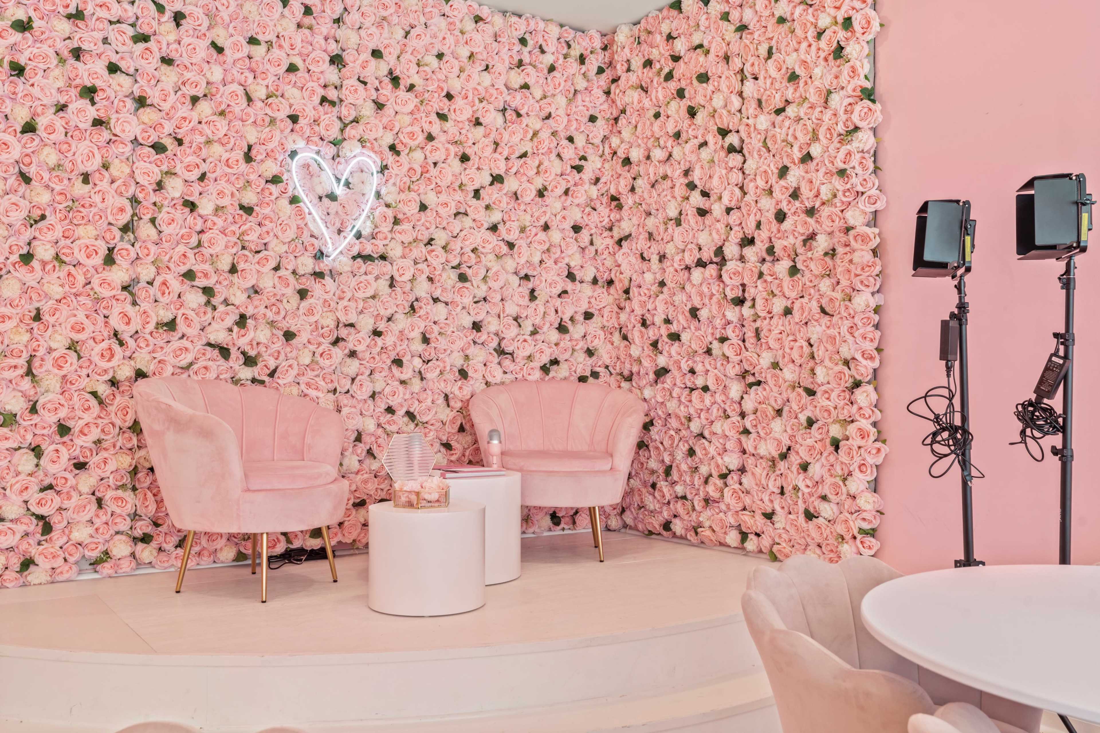A floral backdrop made of pink roses, with two pink velvet chairs and a white table in a well-lit space.