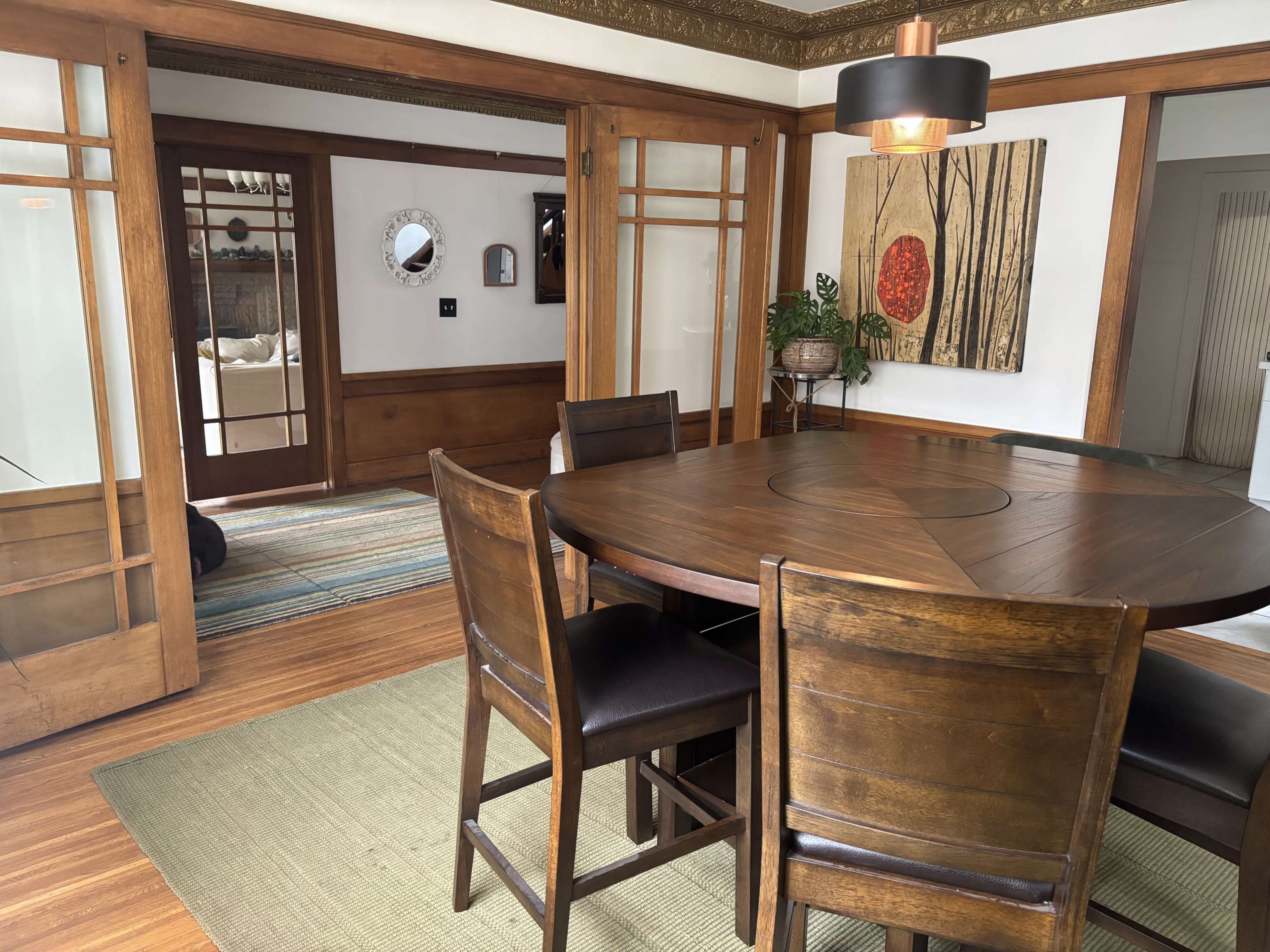 The image shows a dining area with a wooden round table and dark chairs, flanked by sliding glass doors leading to another room and decorated with wall art and a plant.