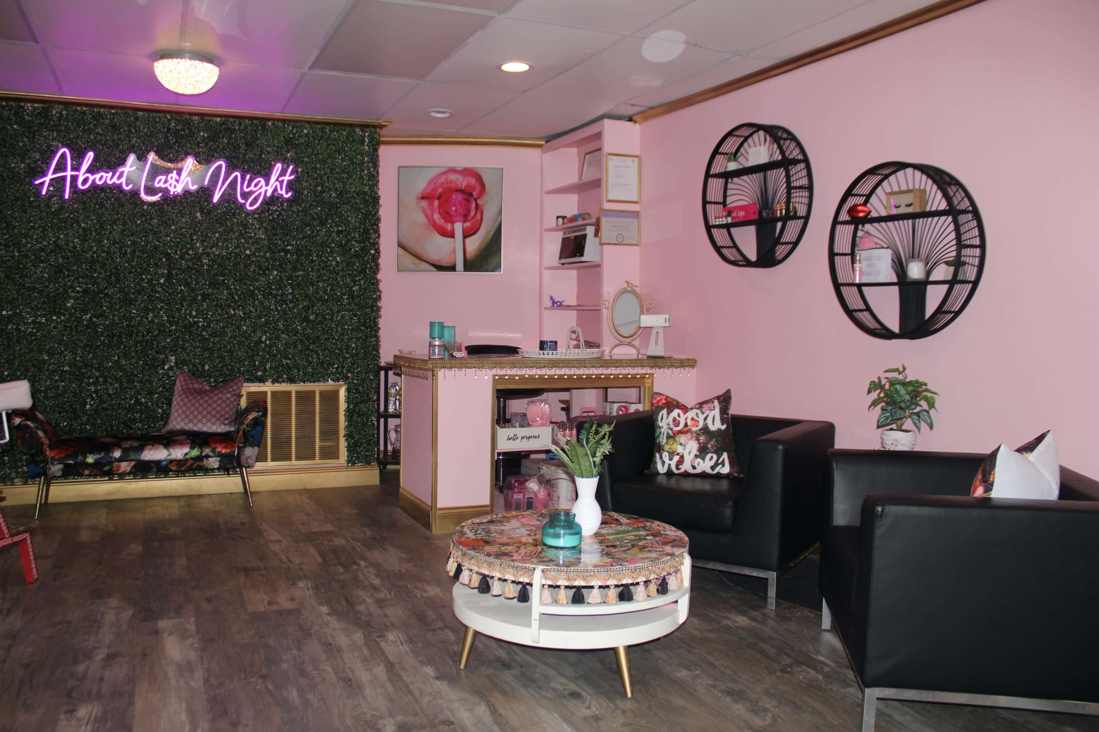 A modern beauty salon interior with pink walls, a neon sign that reads "About Lash Night," and a waiting area featuring two black chairs, a decorative coffee table, and wall decor.