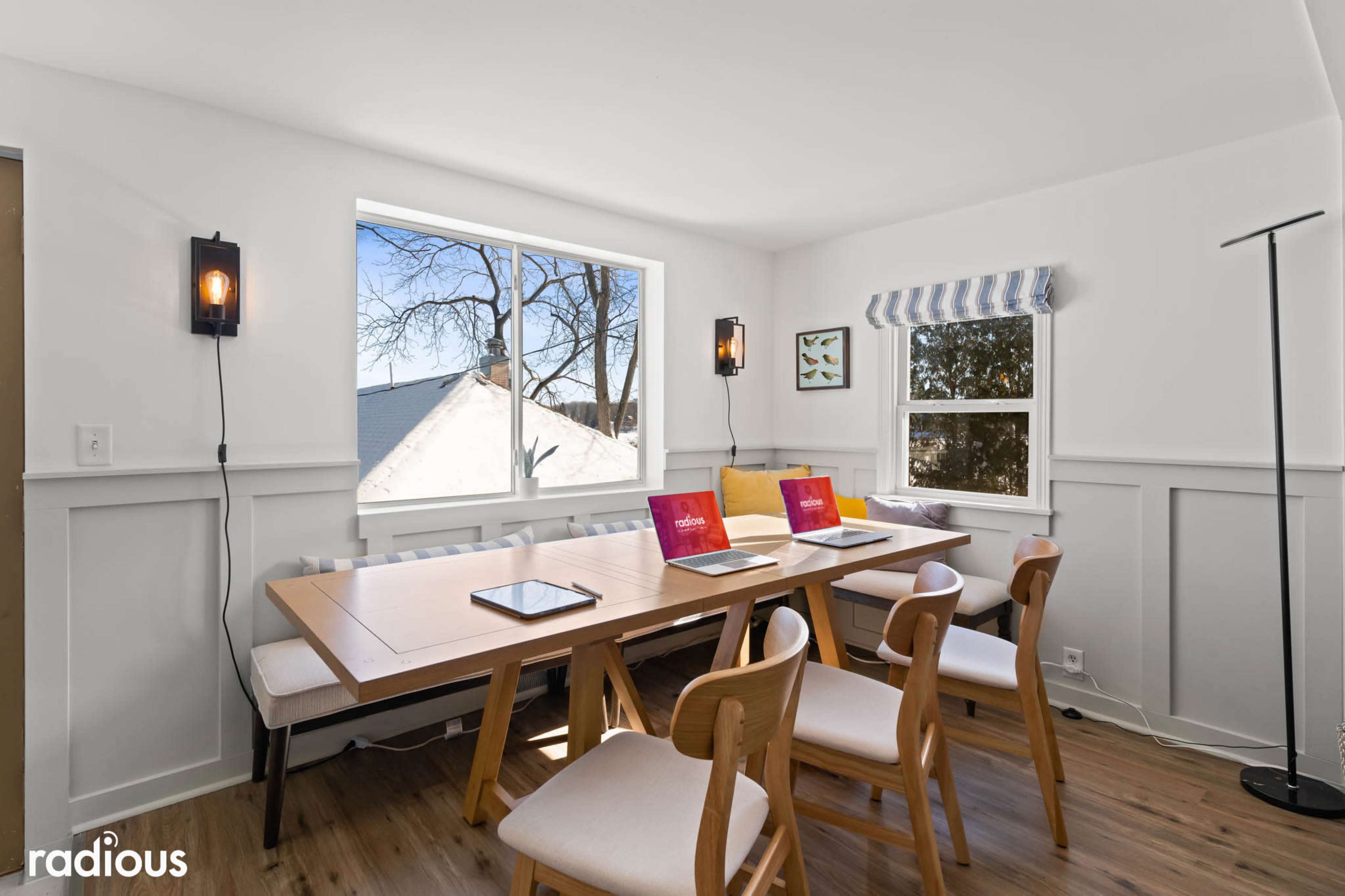 A brightly lit dining area features a long wooden table with two laptops and several chairs, set beside a large window that provides an outside view.
