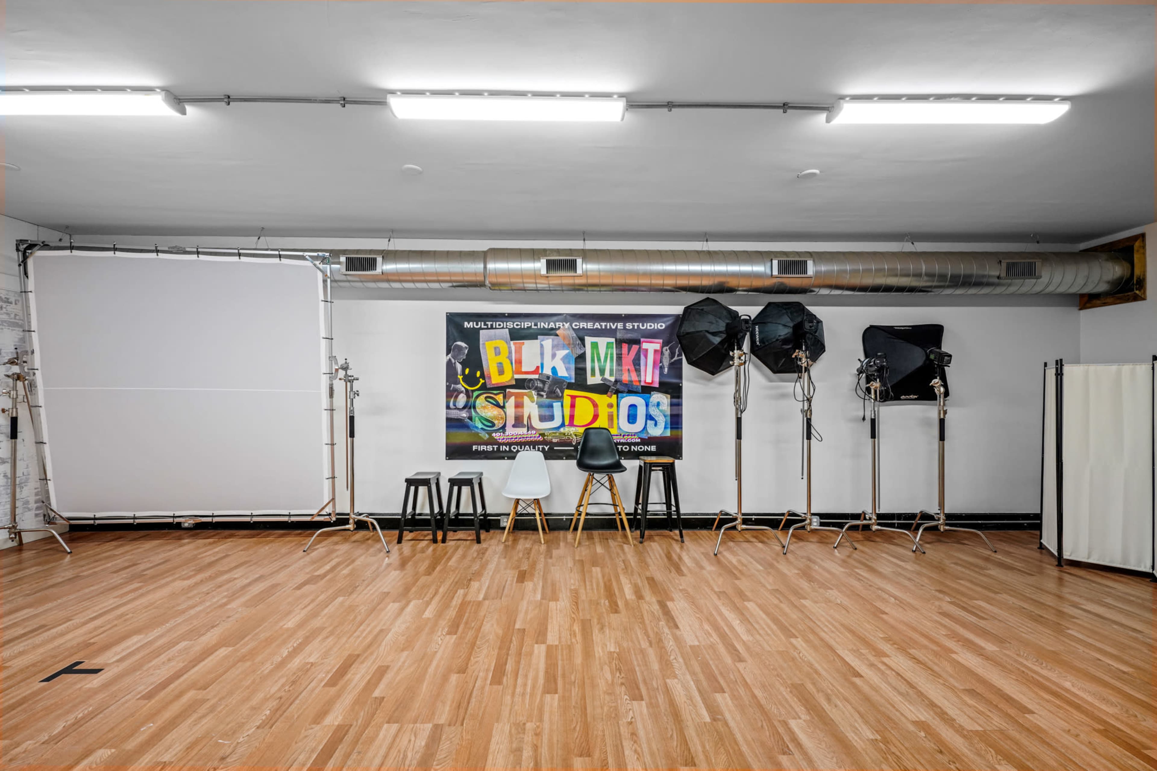 A photography studio featuring a backdrop with "BLK MKT STUDIOS," several lighting setup stands, and a few chairs on a wooden floor.
