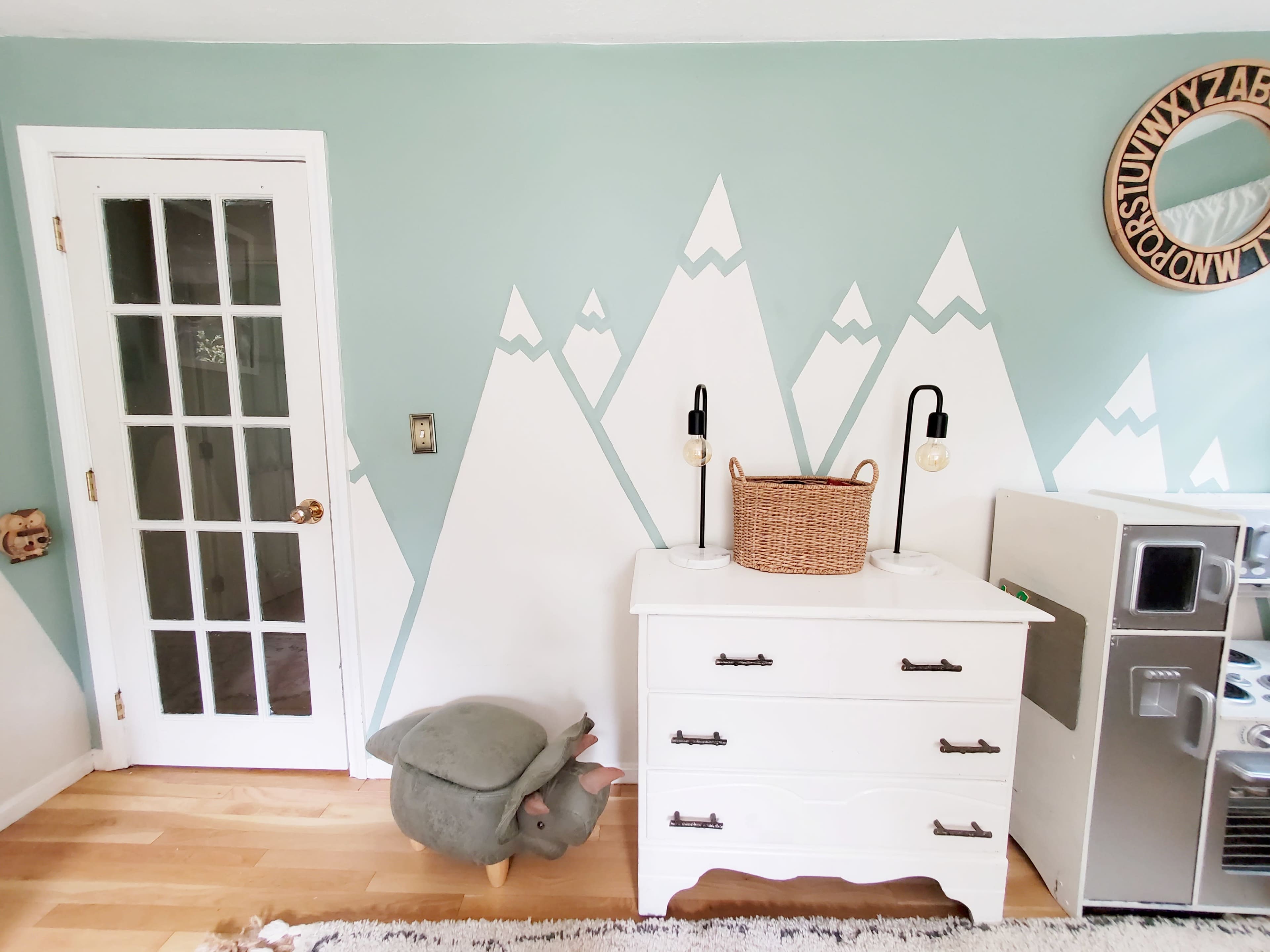 A room with light green walls featuring mountain-shaped designs, a white dresser with handles, a wicker basket on top, a turtle-shaped stool, and a glass panel door.