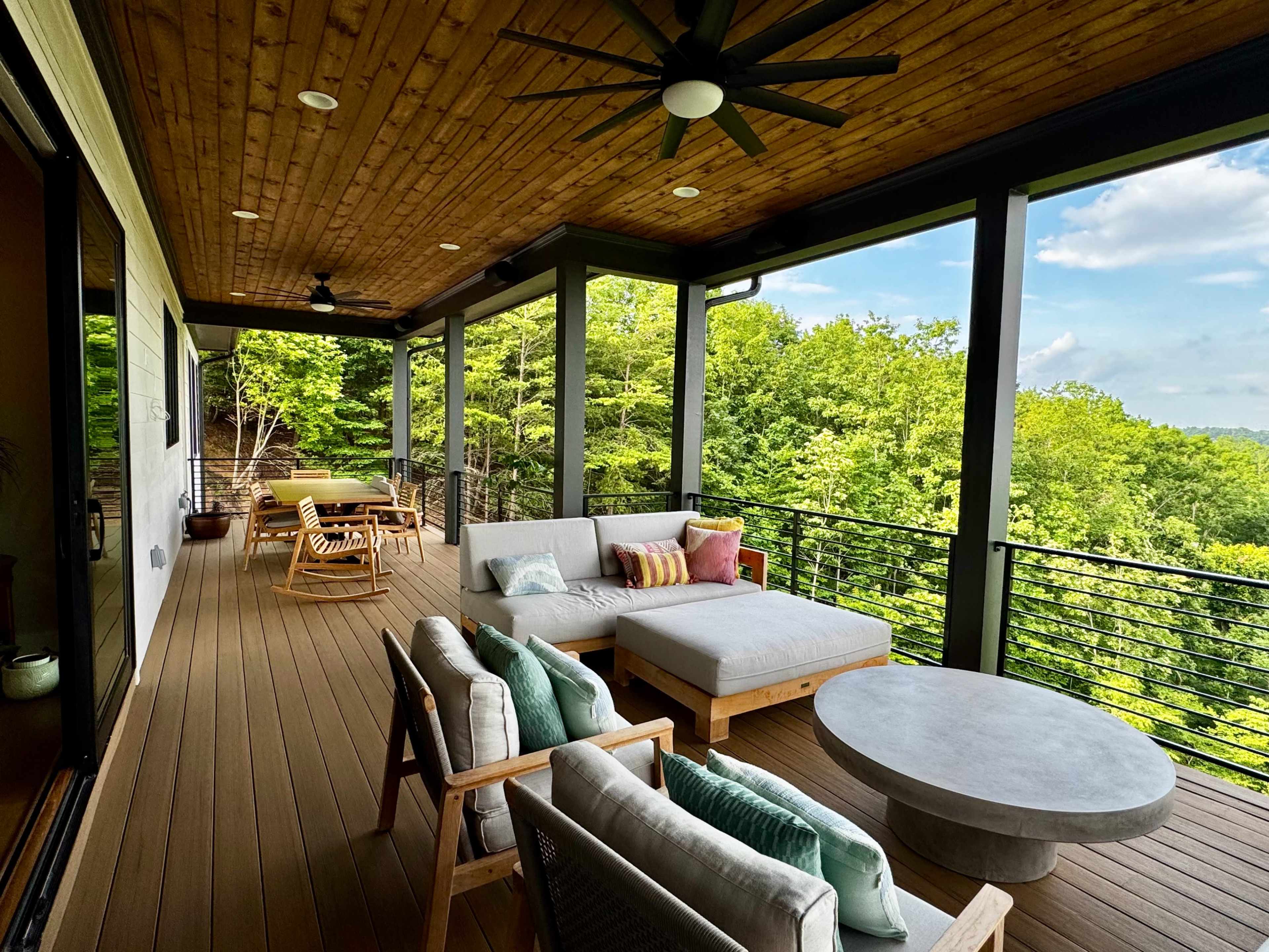 The image shows a spacious outdoor patio featuring comfortable seating, a coffee table, and a dining area, surrounded by trees and open sky.