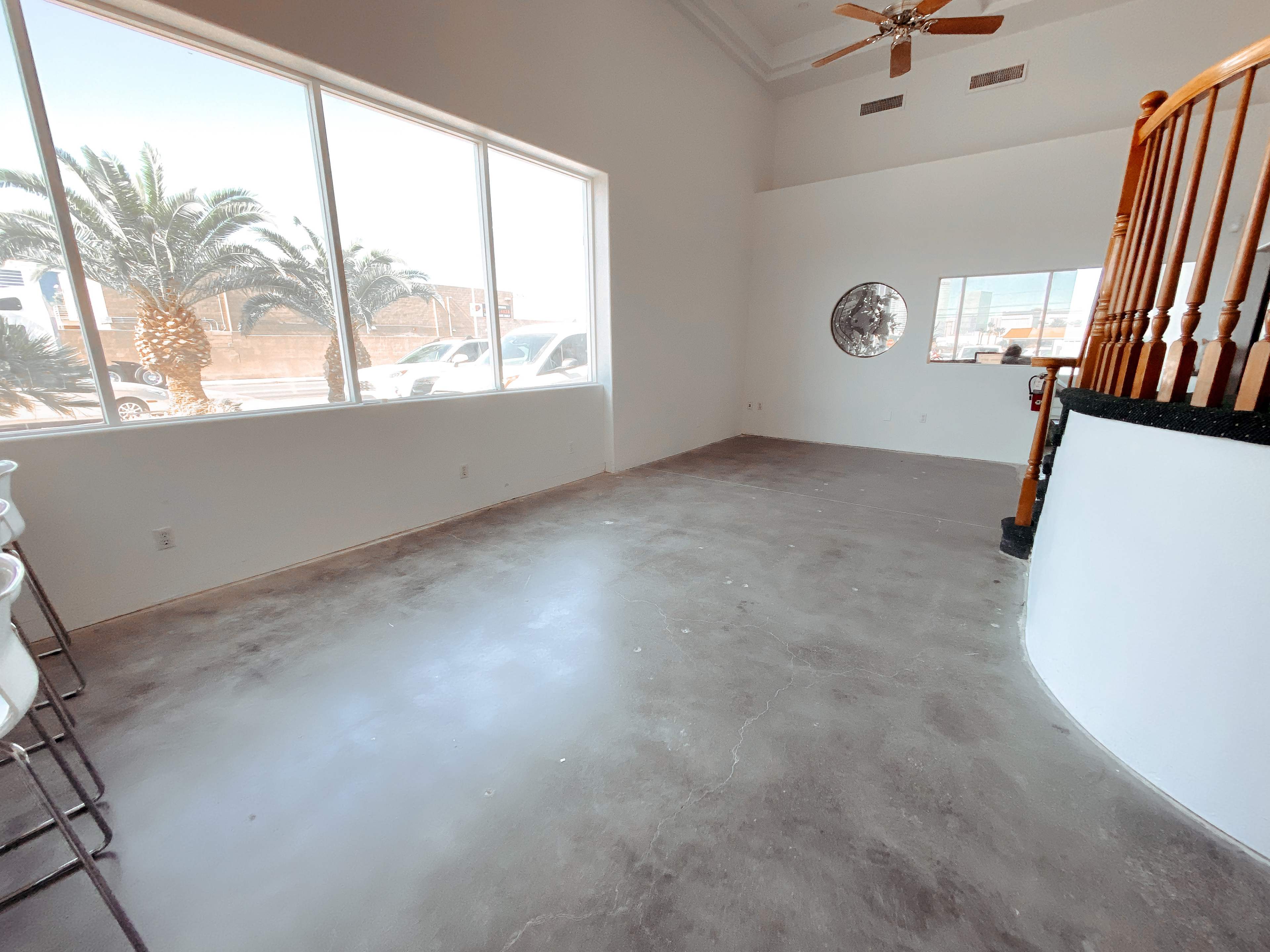 A spacious, empty room with large windows, a curved staircase, and concrete flooring.