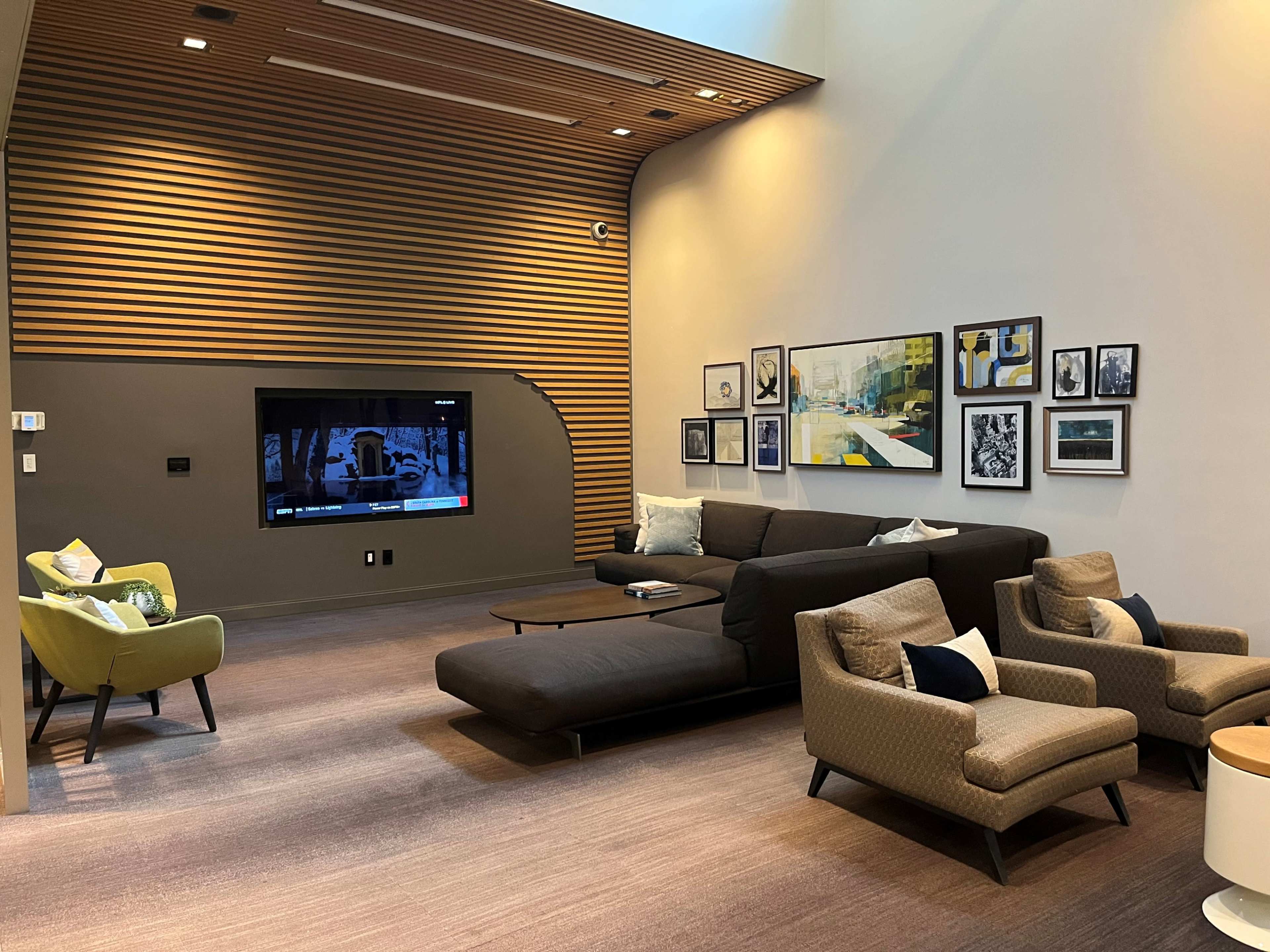 The image shows a modern living room with a sectional sofa, two armchairs, a television mounted on a wooden wall, and several framed artworks arranged around the space.