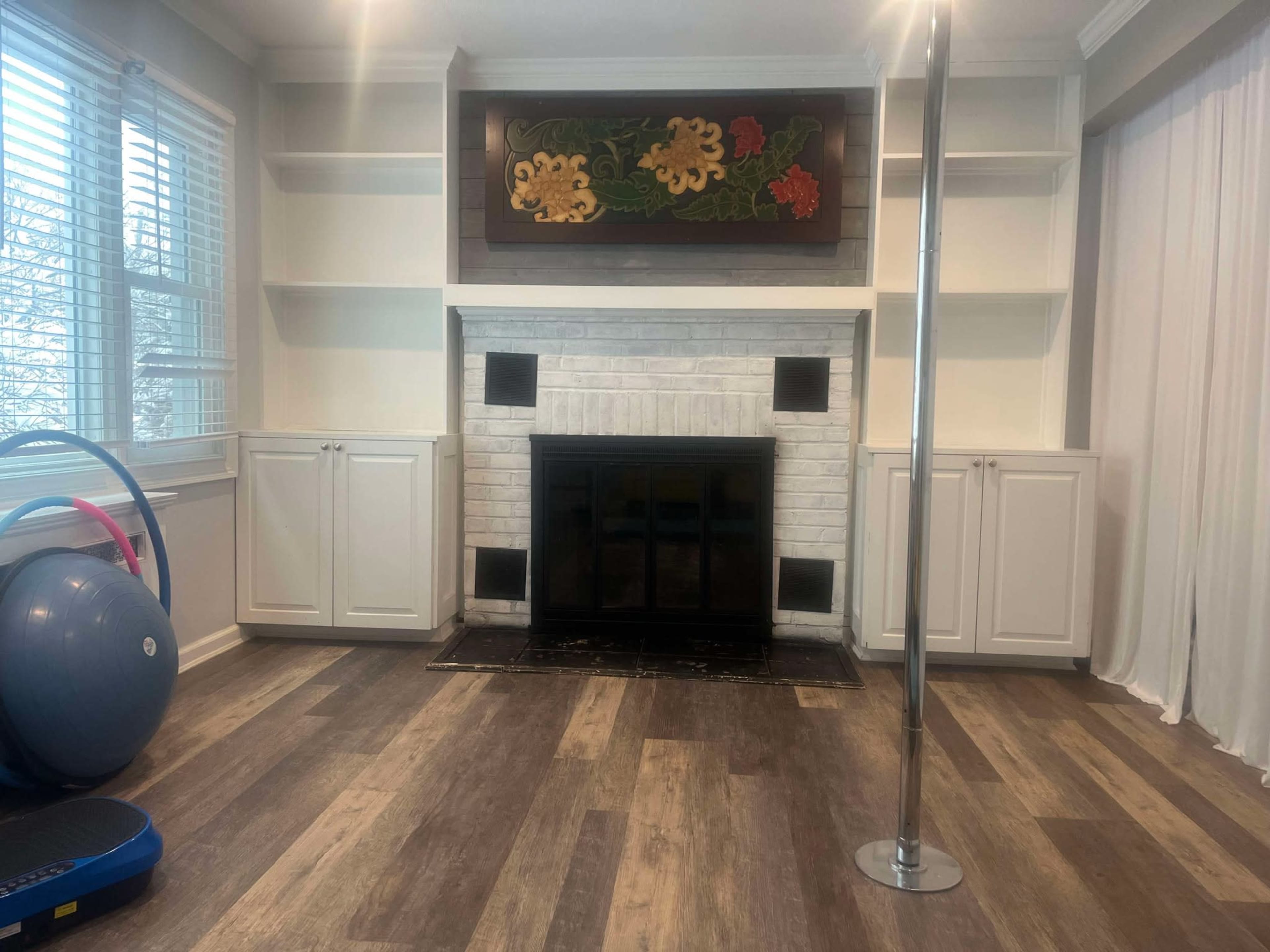 The image shows a room featuring a fireplace with a white brick surround, built-in shelves, a decorative wall piece, a pole, and exercise equipment, all placed on a wooden floor.