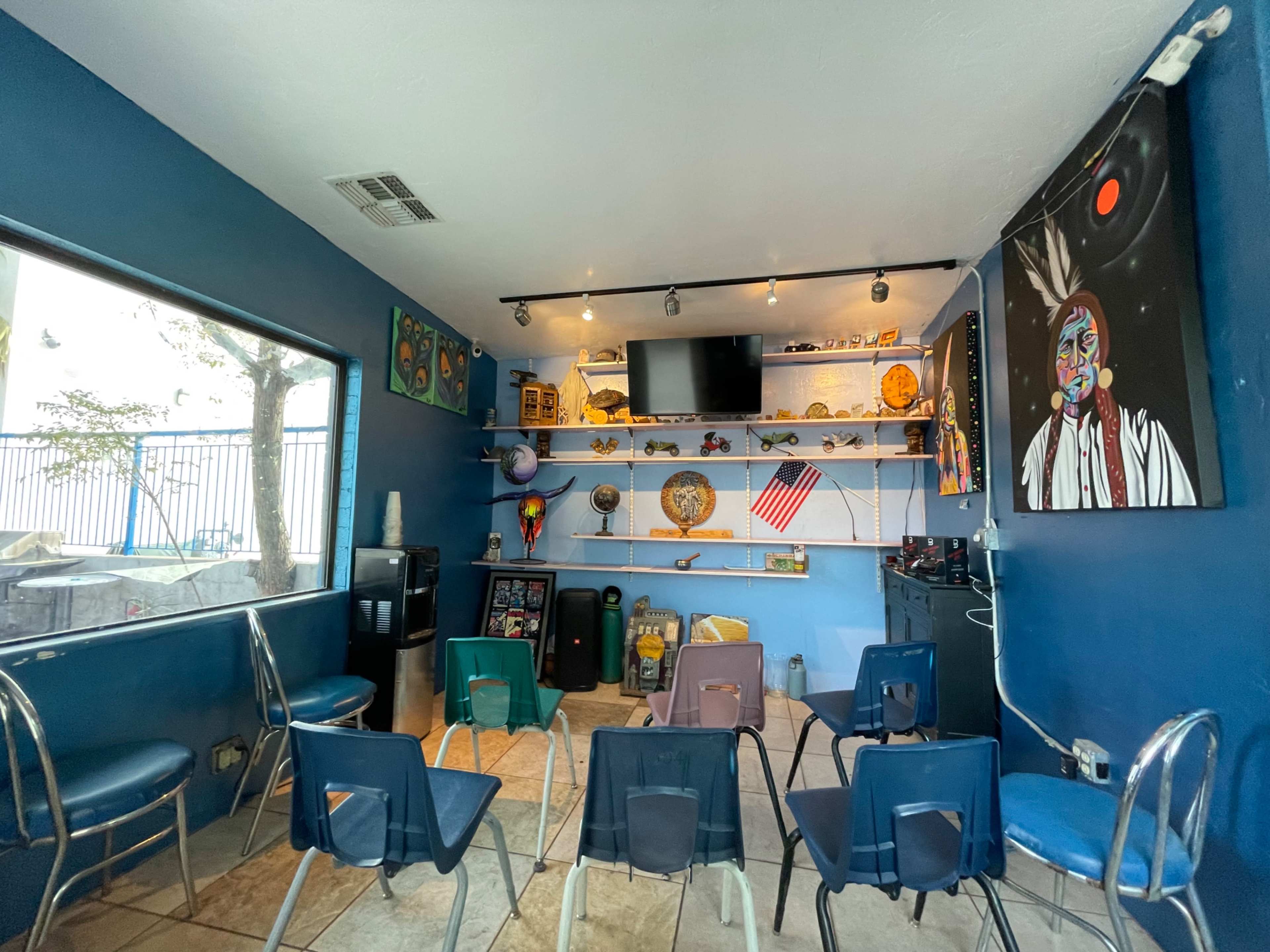 The image shows a small room with blue walls, several shelves displaying various objects, a TV mounted on the wall, and a row of empty folding chairs arranged in a semicircle.