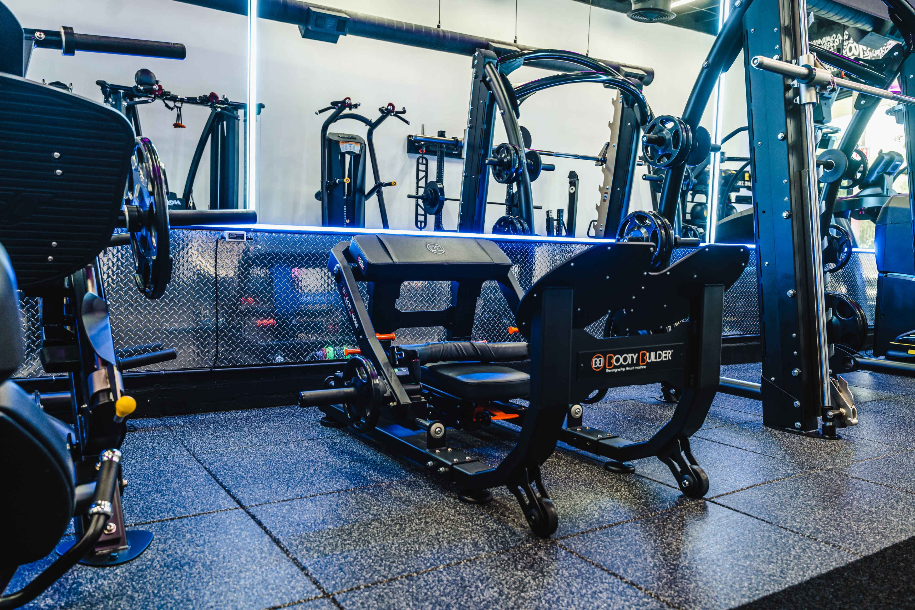 The image features a modern gym interior with various fitness equipment, including a black and orange weight machine prominently in the foreground.