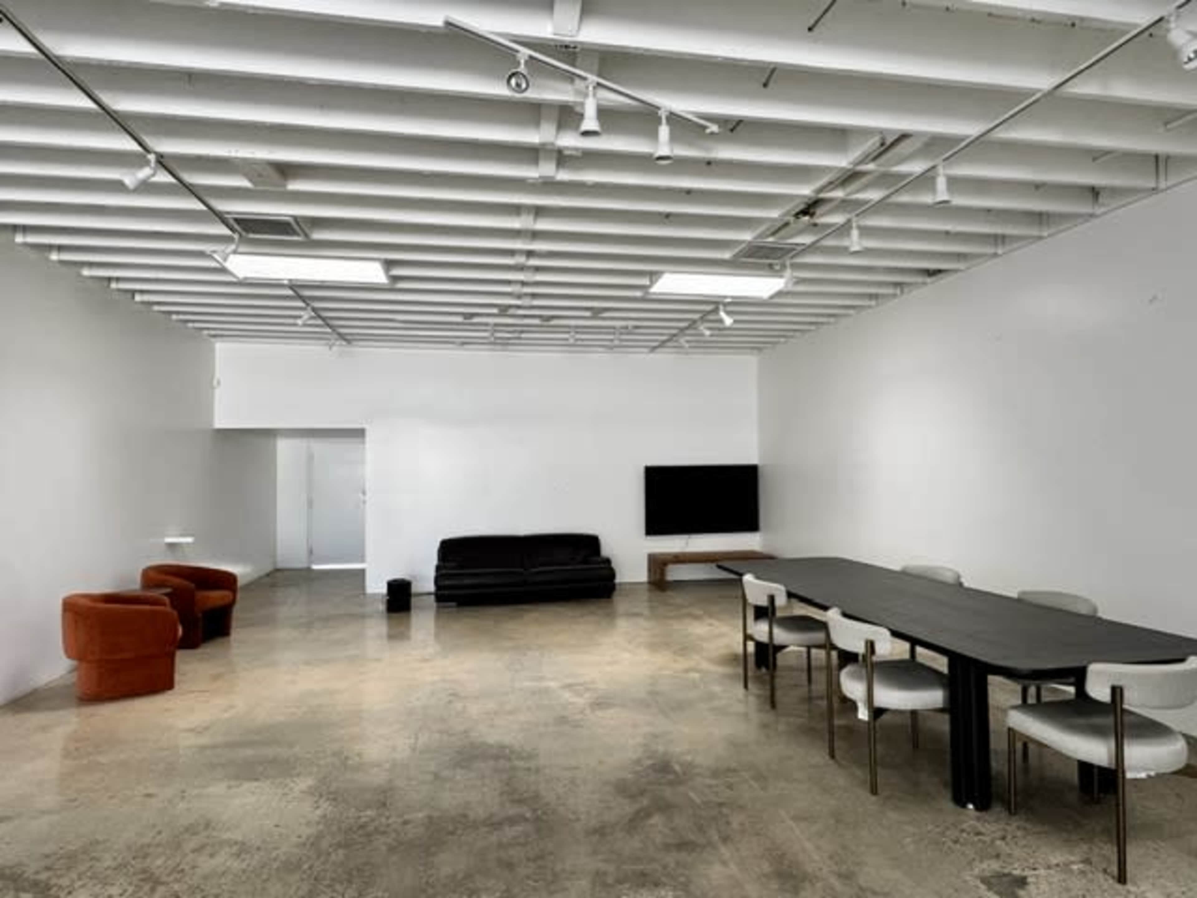 The image shows a spacious, minimalistic room with a black sofa, two orange chairs, a long table with white chairs, and a television against a white wall.