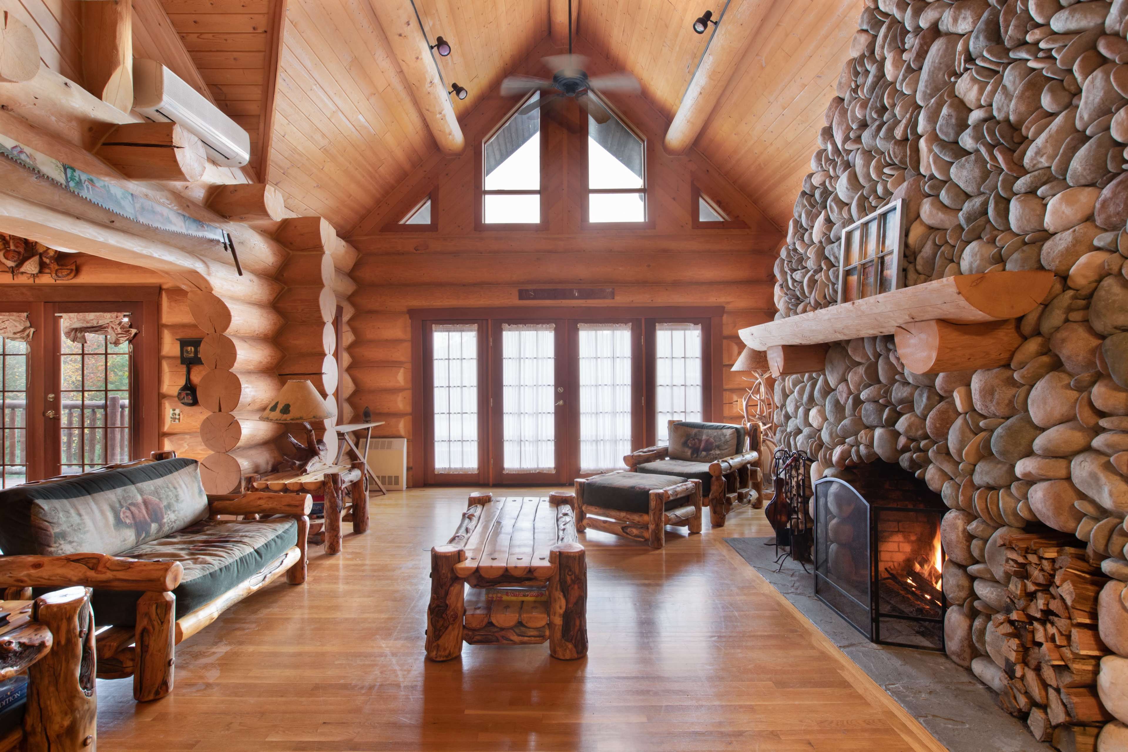 The interior of a log cabin features a stone fireplace, wooden furniture, and large windows that provide natural light.