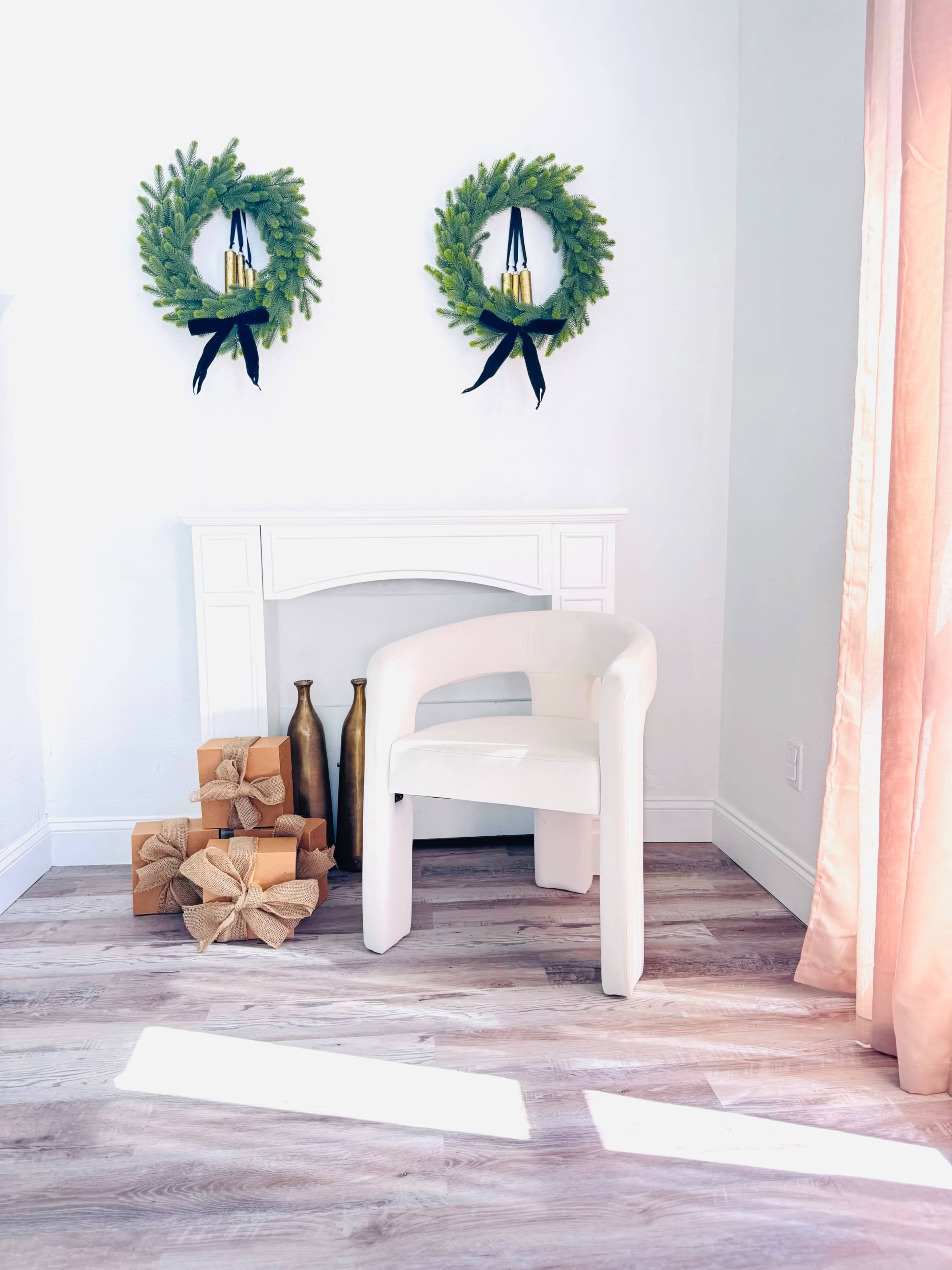 A white chair is positioned near a small fireplace, with two green wreaths hanging above and wrapped gifts stacked beside gold vases.