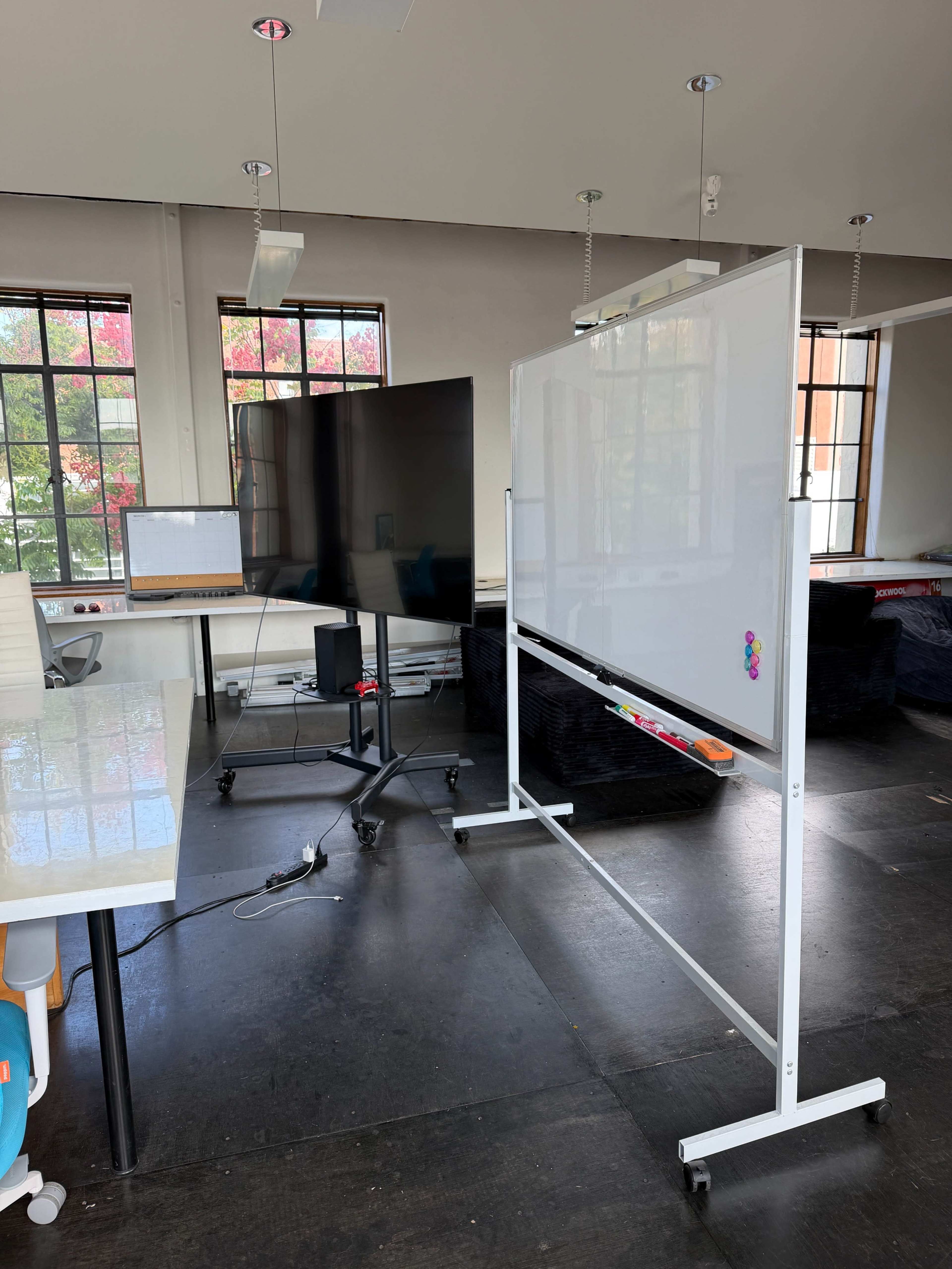 The image shows a bright room featuring a whiteboard on a stand next to a large screen and a computer setup on a table.