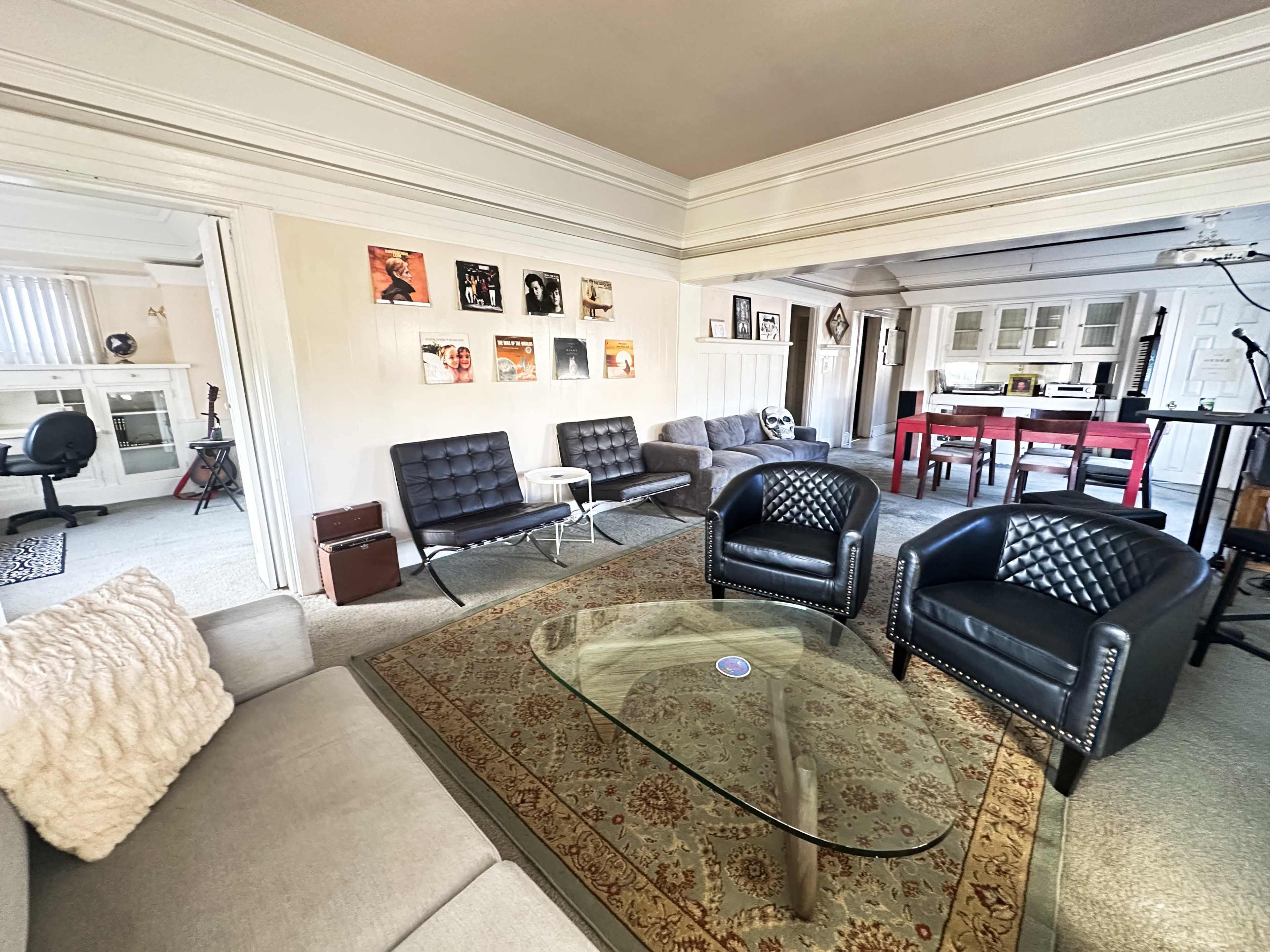 The image shows a spacious living area with various seating options, including a gray sofa, black armchairs, and a glass coffee table, contrasting with a red dining table visible in the adjacent kitchen area.