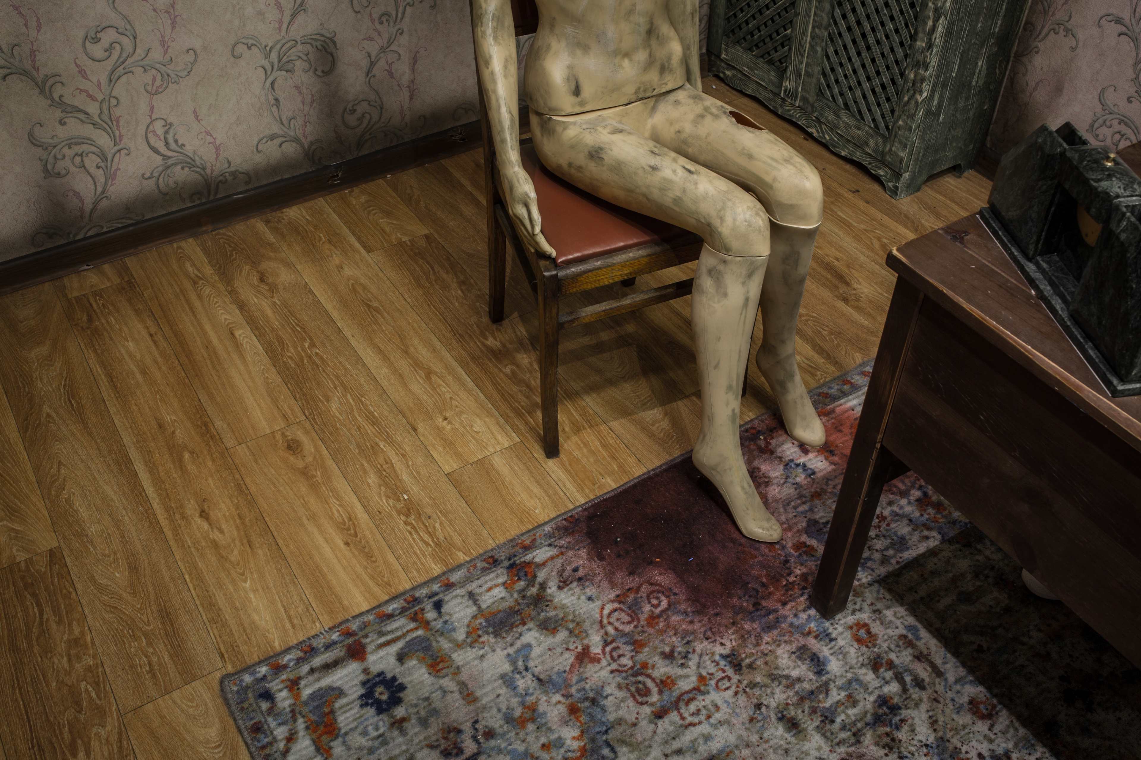 A partially dressed mannequin sits on a wooden chair beside a table in a room with patterned wallpaper and a stained rug.
