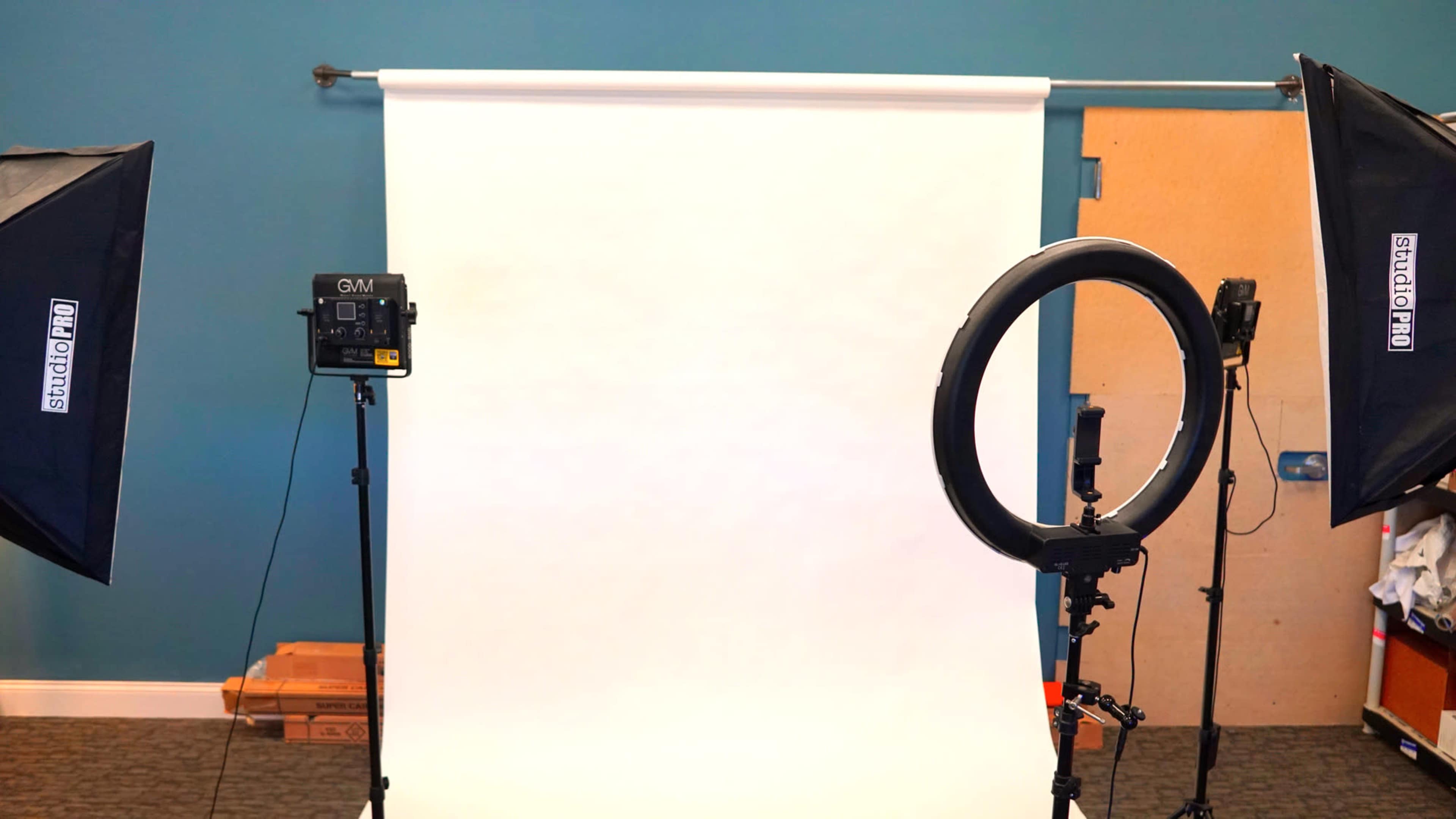 The image shows a photography studio setup with two softbox lights, a ring light, and a blank backdrop.