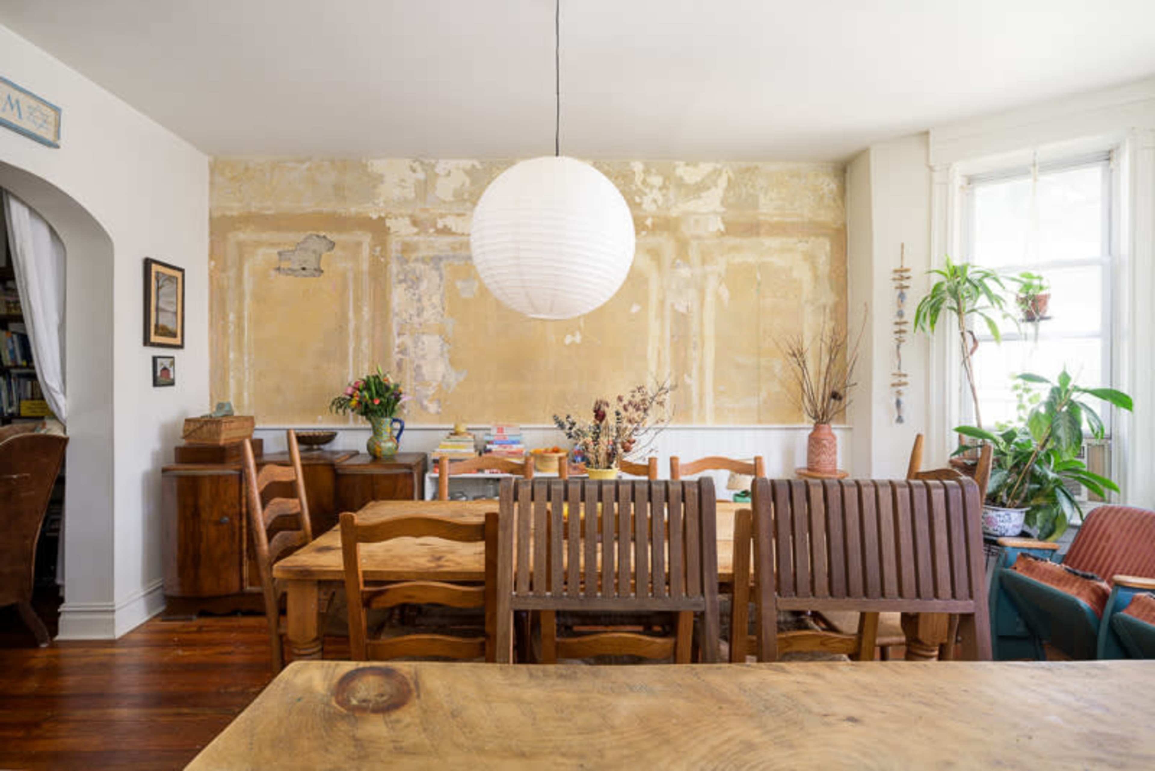 The image shows a rustic dining area with a large wooden table surrounded by chairs, a round pendant light hanging above, and a wall with peeling yellow paint adorned with plants and decorative objects.