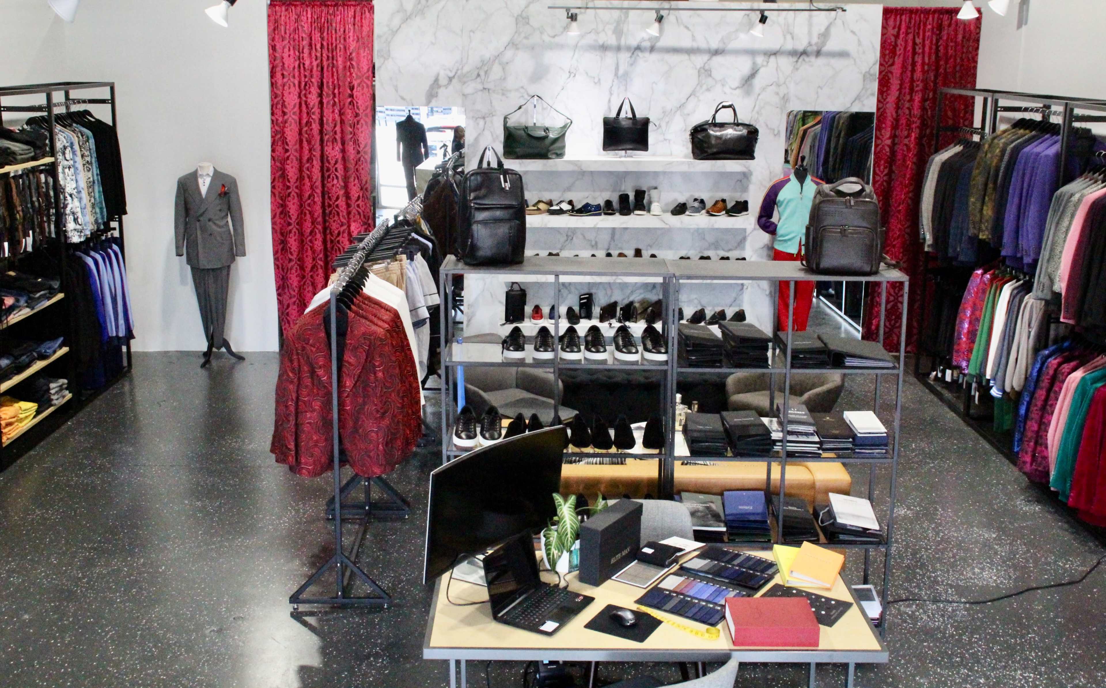 The image shows a retail clothing store featuring various garments displayed on racks, a shelf with shoes, and a desk with assorted merchandise and a computer setup.