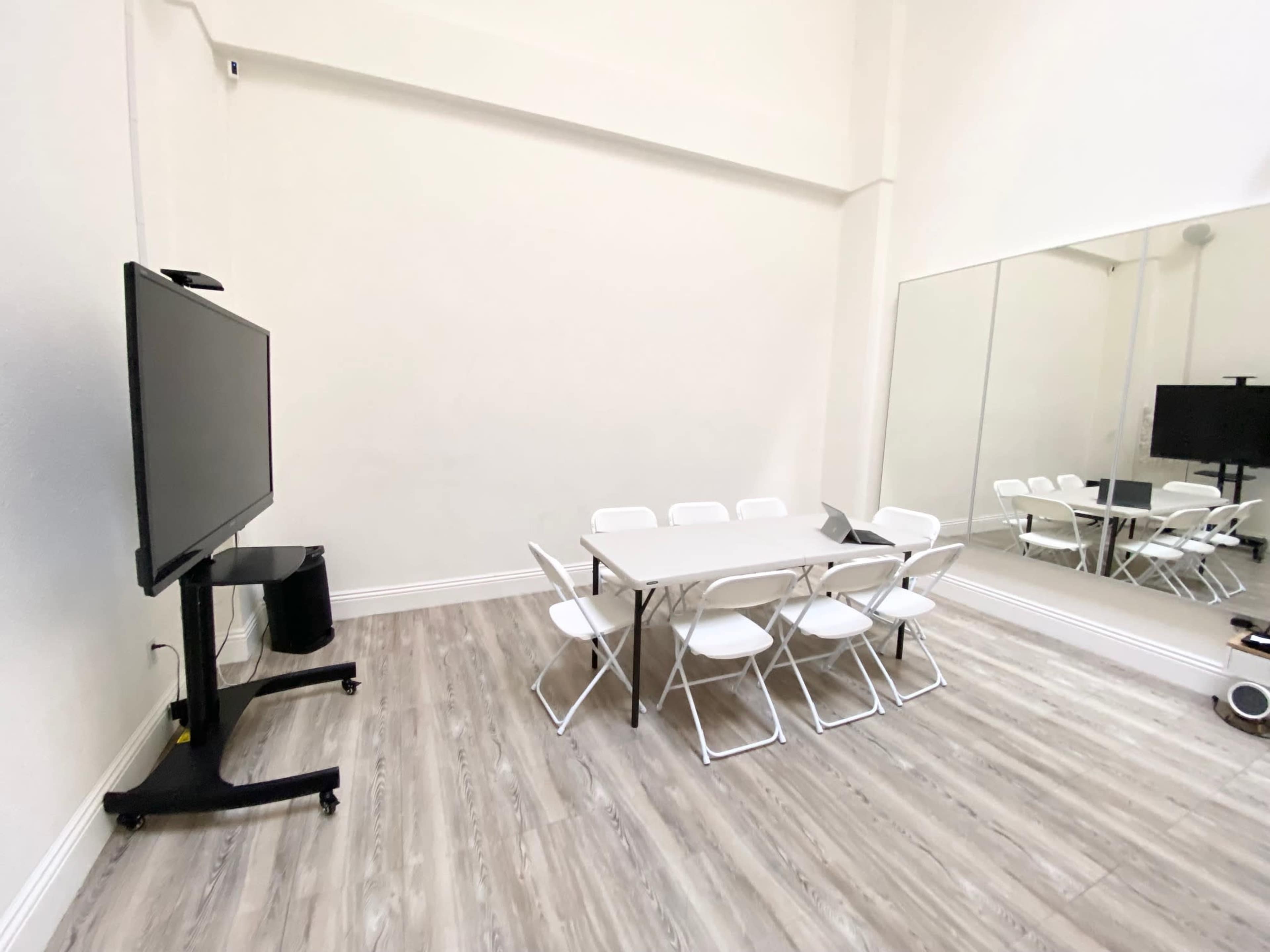 The image depicts a bright meeting room with a large flat-screen TV, a rectangular table surrounded by folding chairs, and a mirror on one wall.