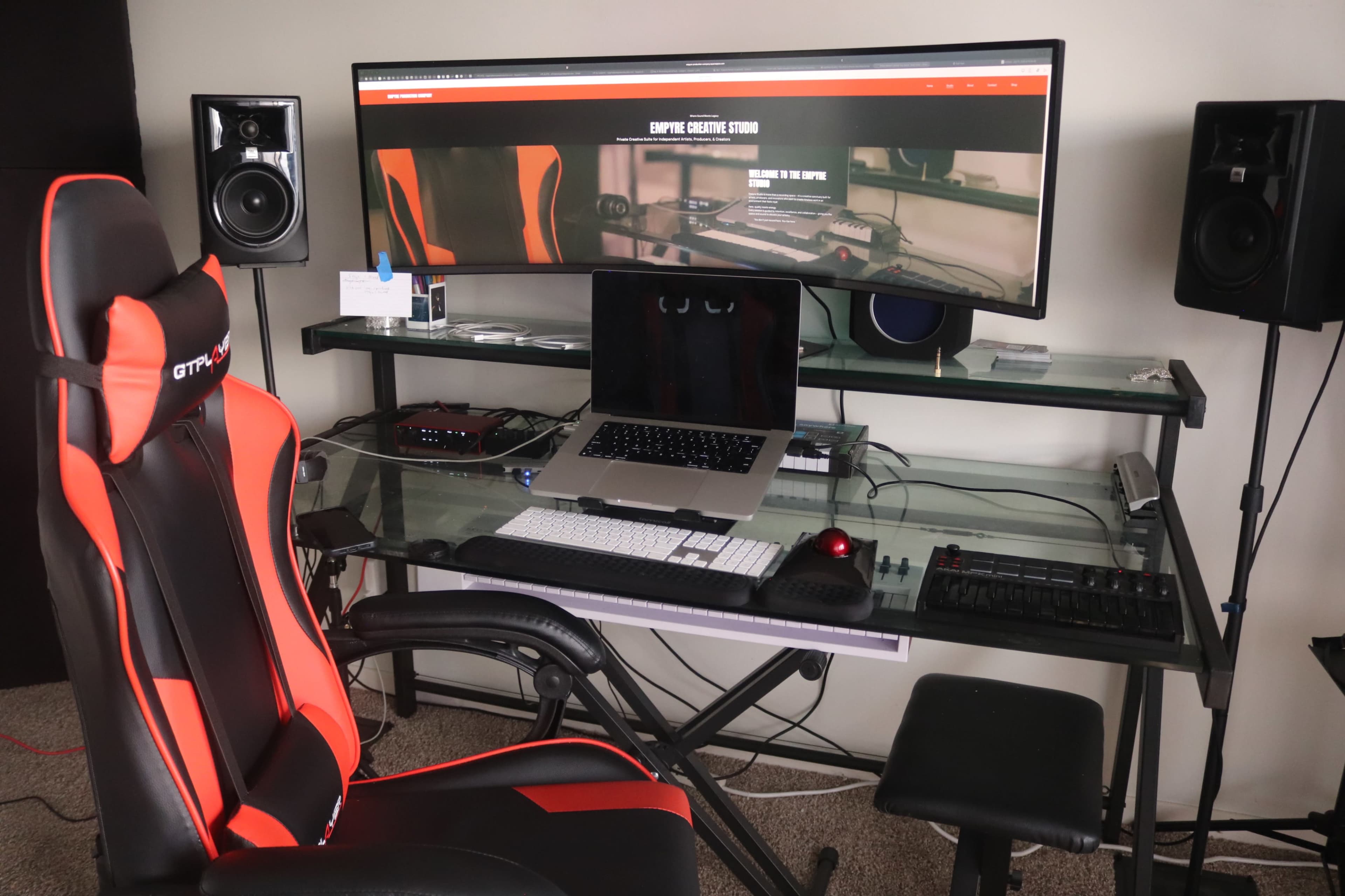 The image shows a modern workspace with an ergonomic gaming chair, a large curved monitor displaying a website, and various computer accessories on a glass desk.