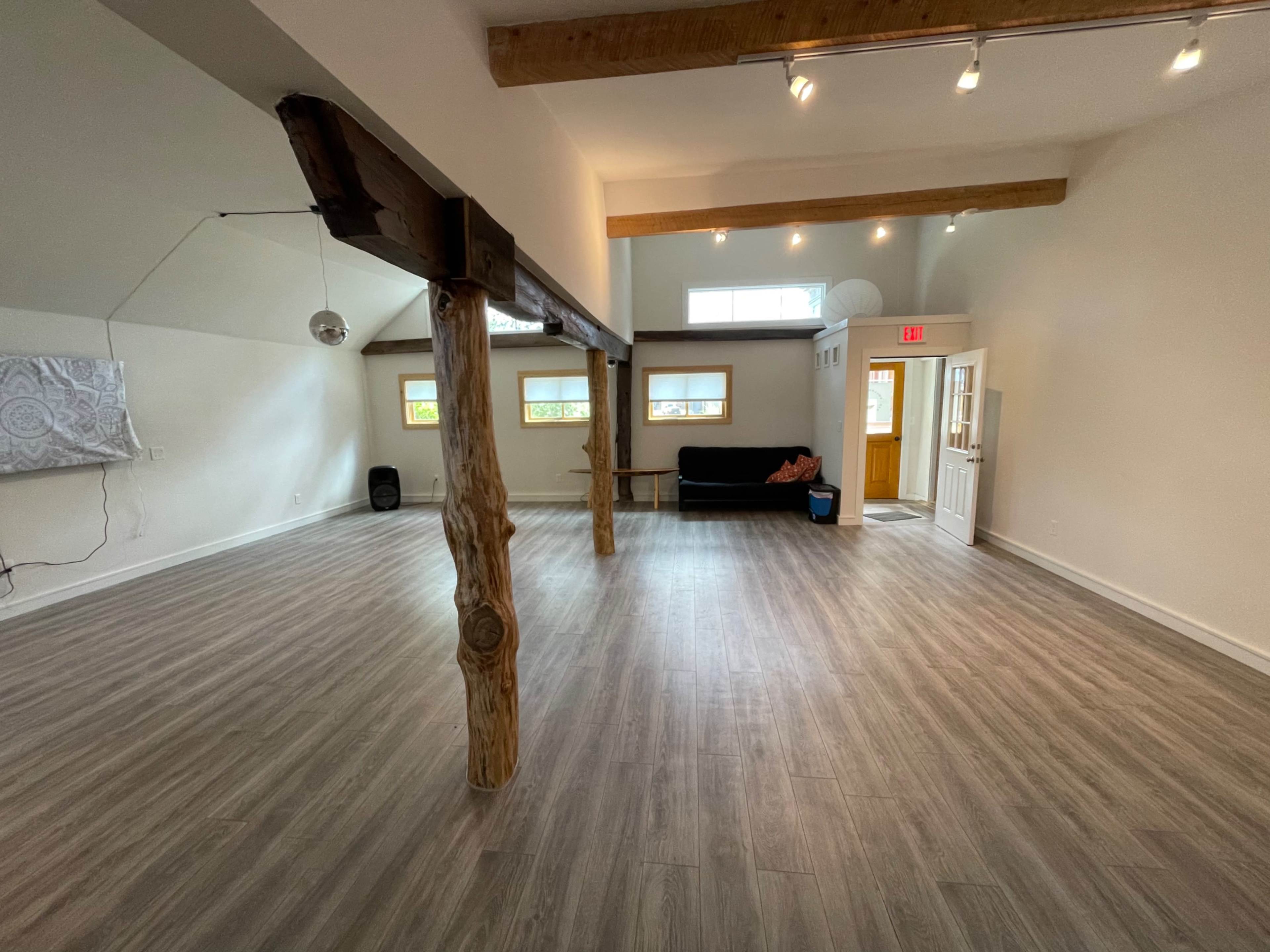 A spacious room with polished wooden floors, exposed wooden beams, and a black couch positioned near the entrance.