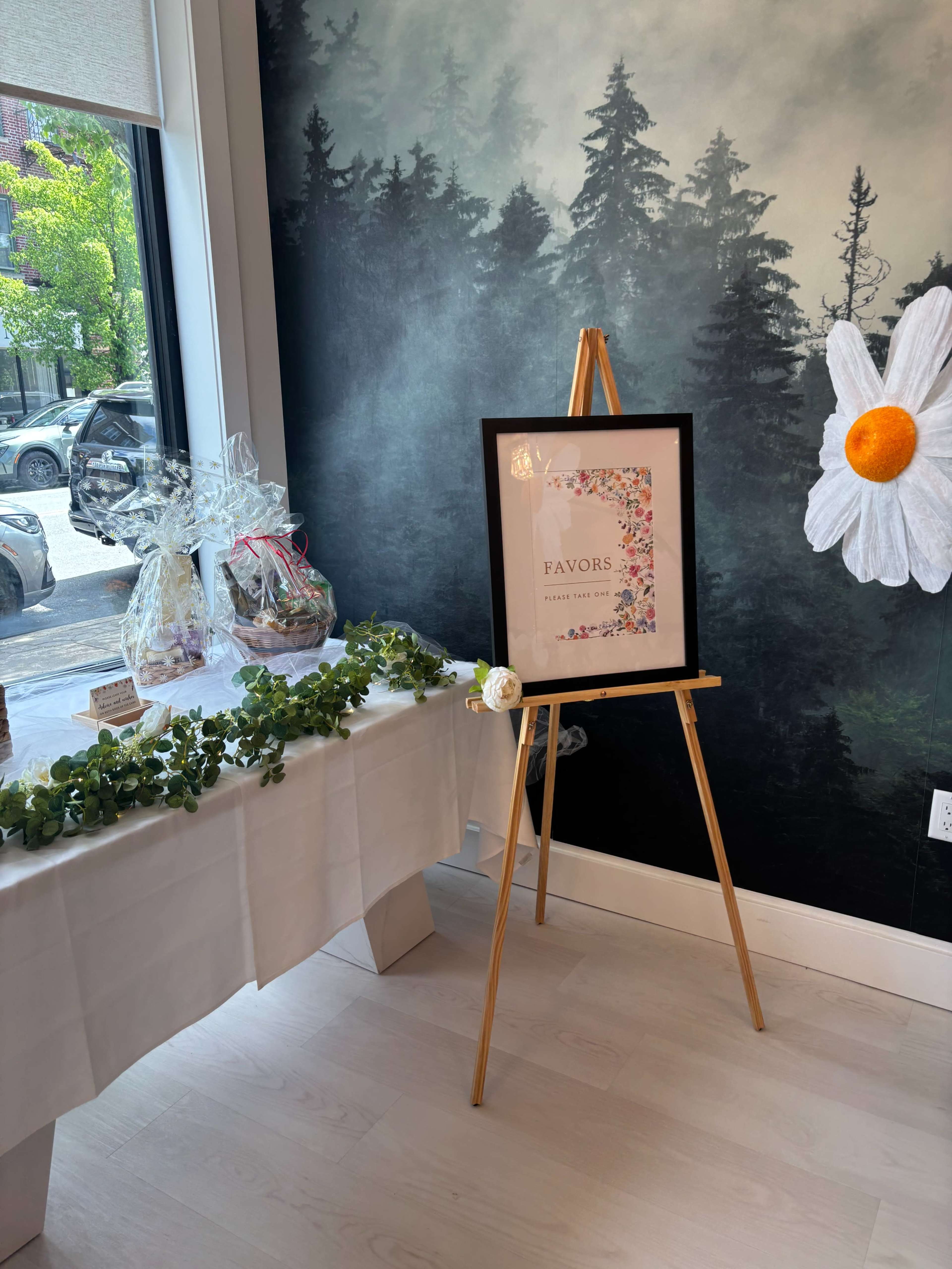 A framed sign reading "FAVORS" is displayed on an easel next to a table decorated with gifts and greenery in a room with a floral mural.