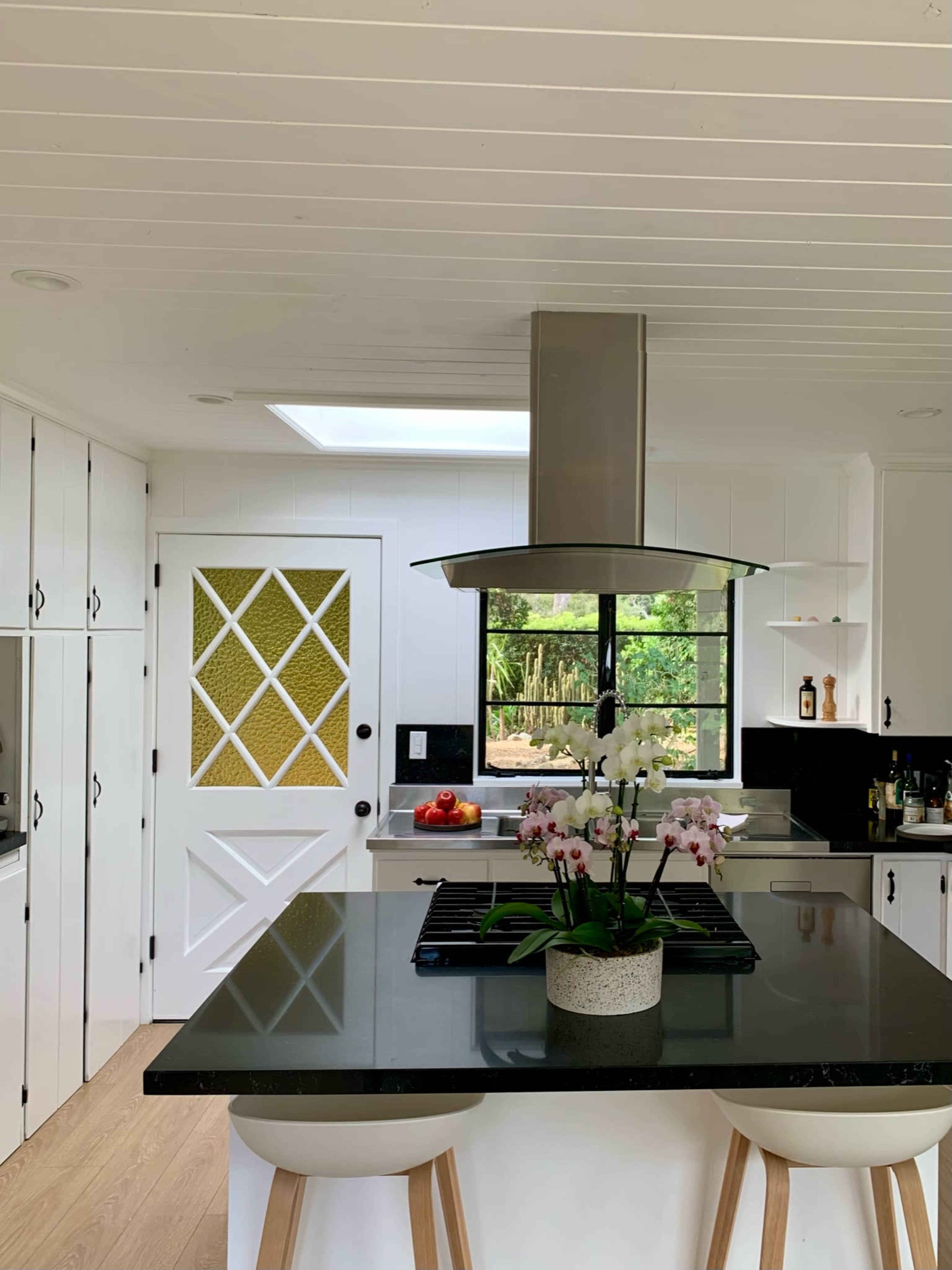 A modern kitchen features a central island with a dark countertop, a hood above the stove, and a large window allowing natural light.