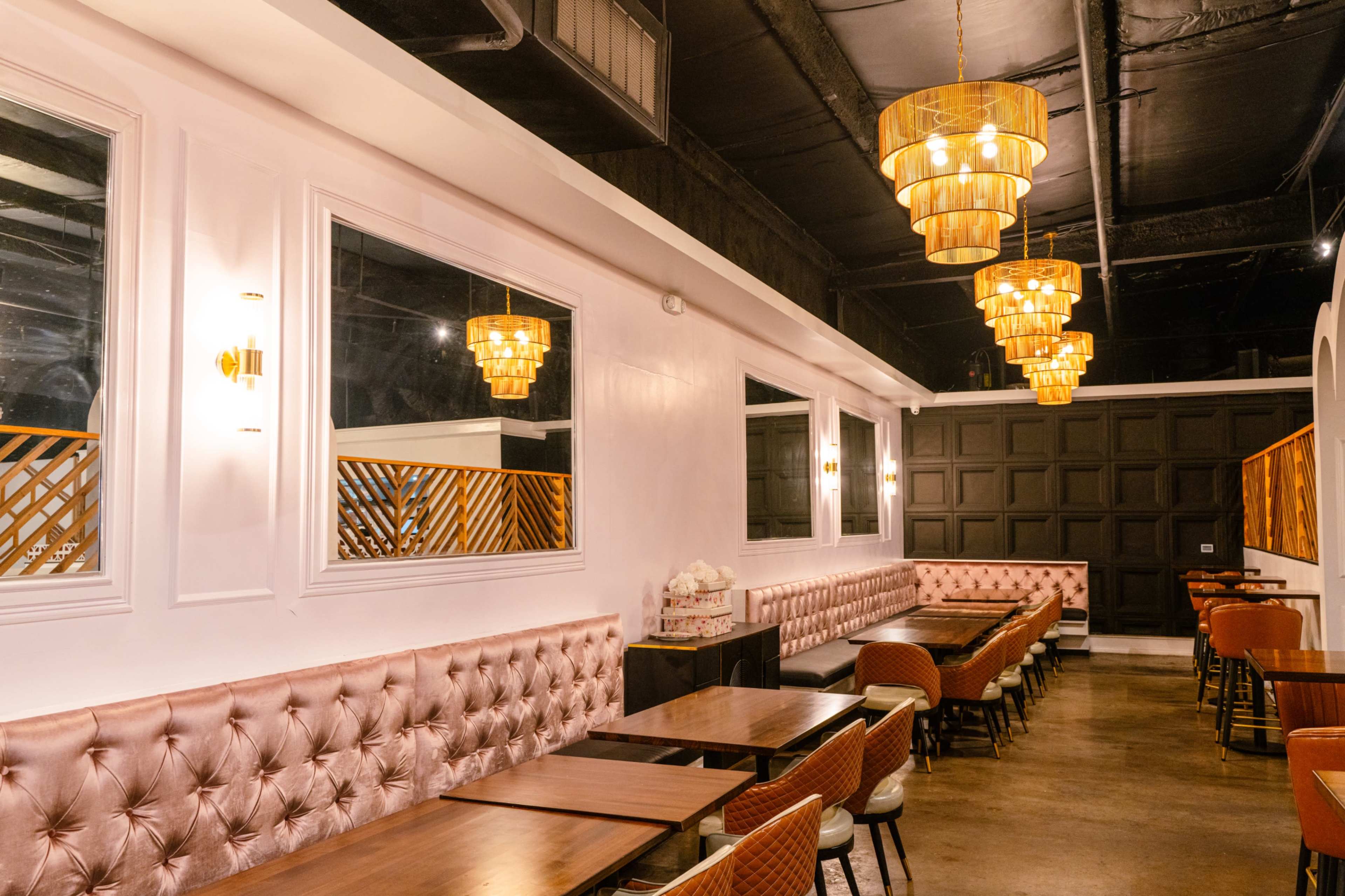 The image depicts a stylish restaurant interior featuring plush pink banquettes, wooden tables, and multiple gold pendant lights, with mirrors and a dark wood accent wall in the background.