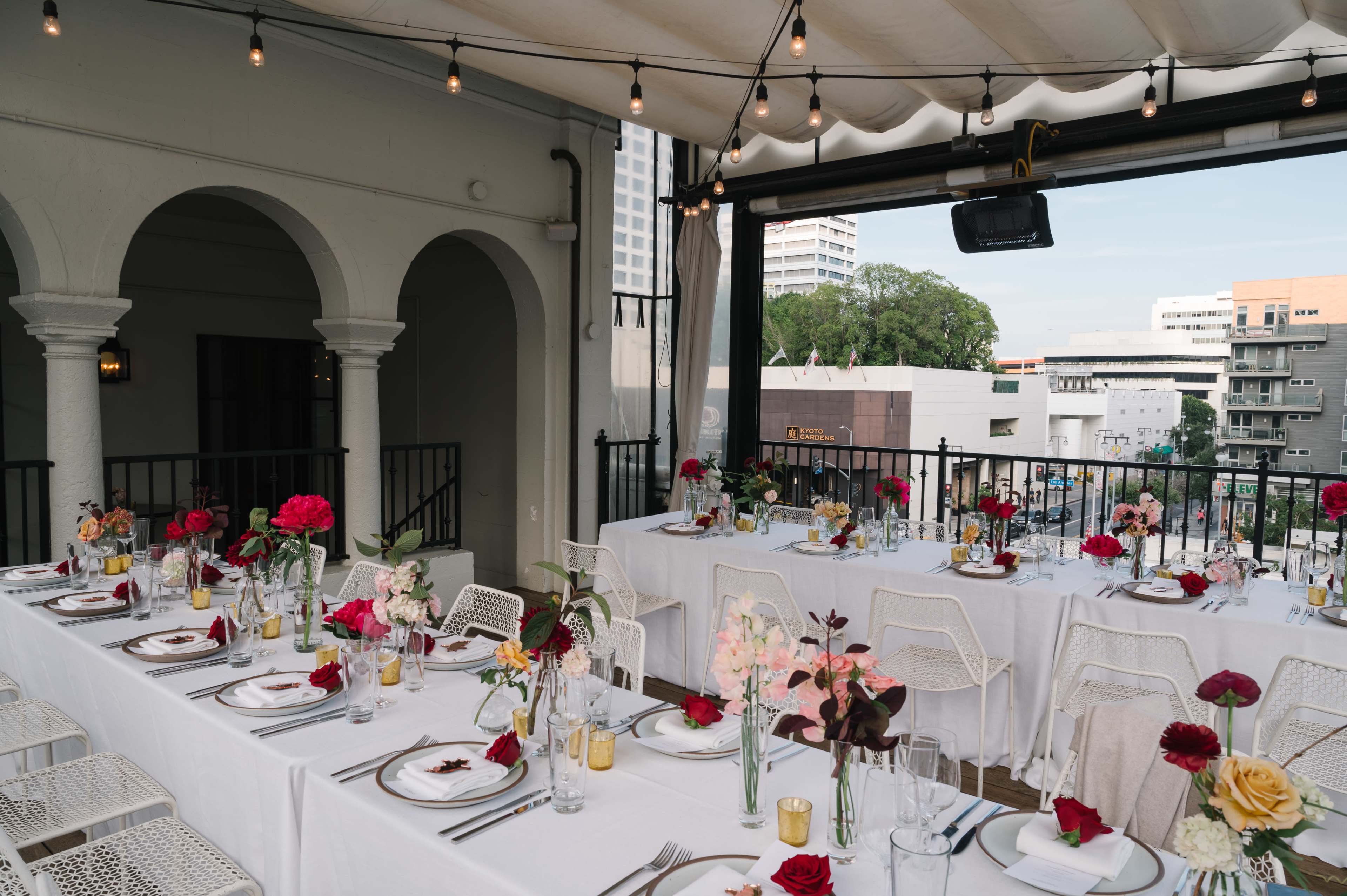 A rooftop dining area features elegantly set tables adorned with flowers and candles, overlooking a cityscape.