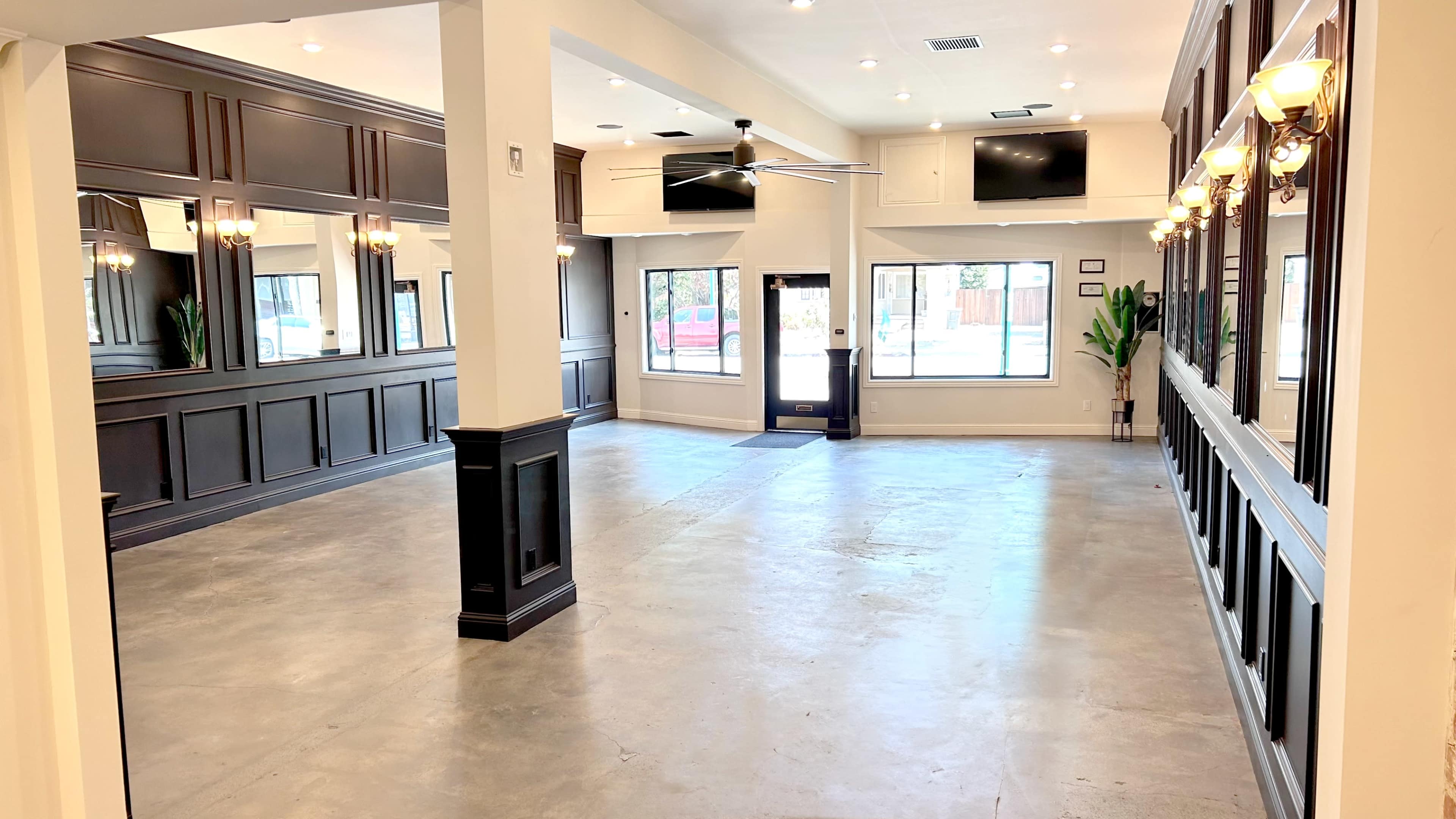 A spacious, well-lit interior room with concrete flooring, large windows, and black-paneled walls adorned with light fixtures.
