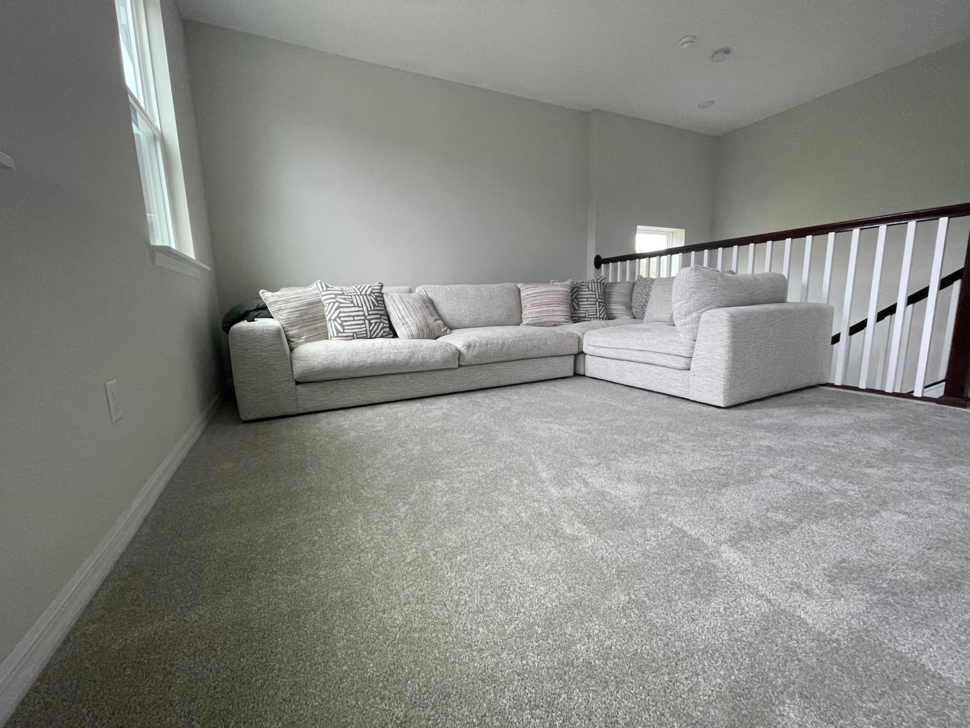 A large sectional sofa with decorative pillows is positioned in a carpeted room with light gray walls and a railing visible in the background.