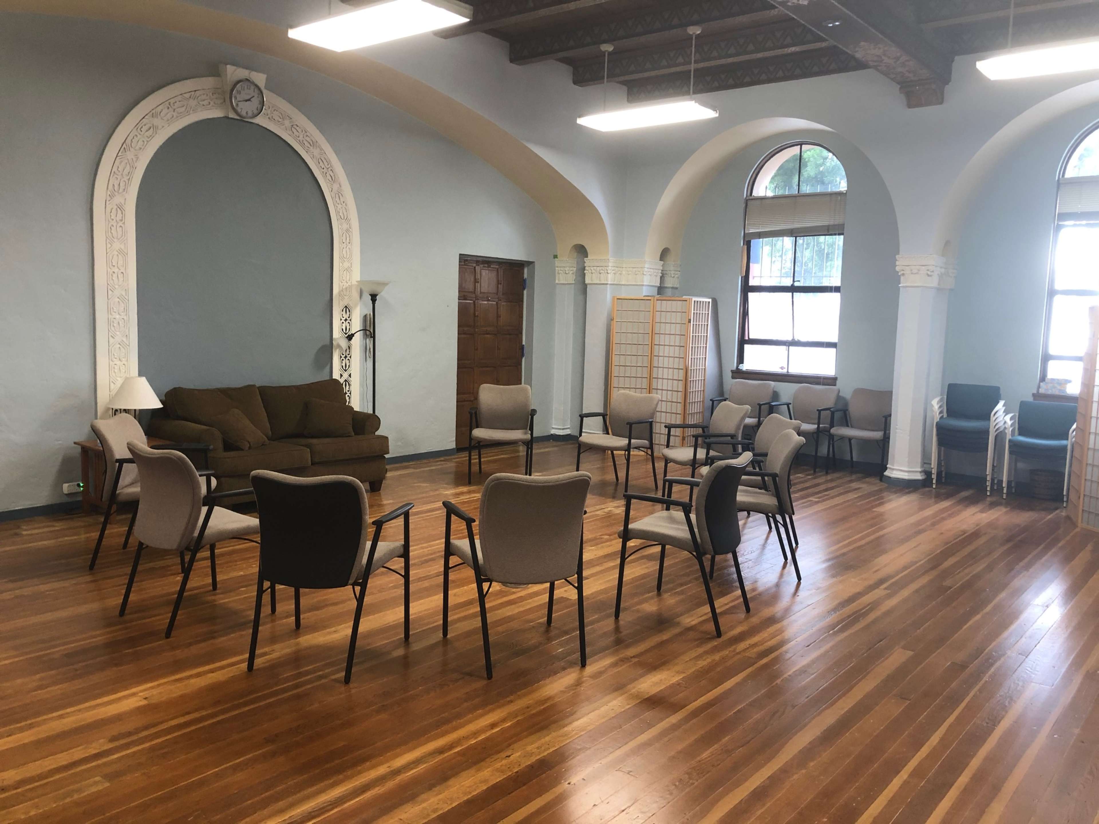 A spacious room with hardwood floors features a circular arrangement of chairs around a sofa, with large windows and decorative architectural details.
