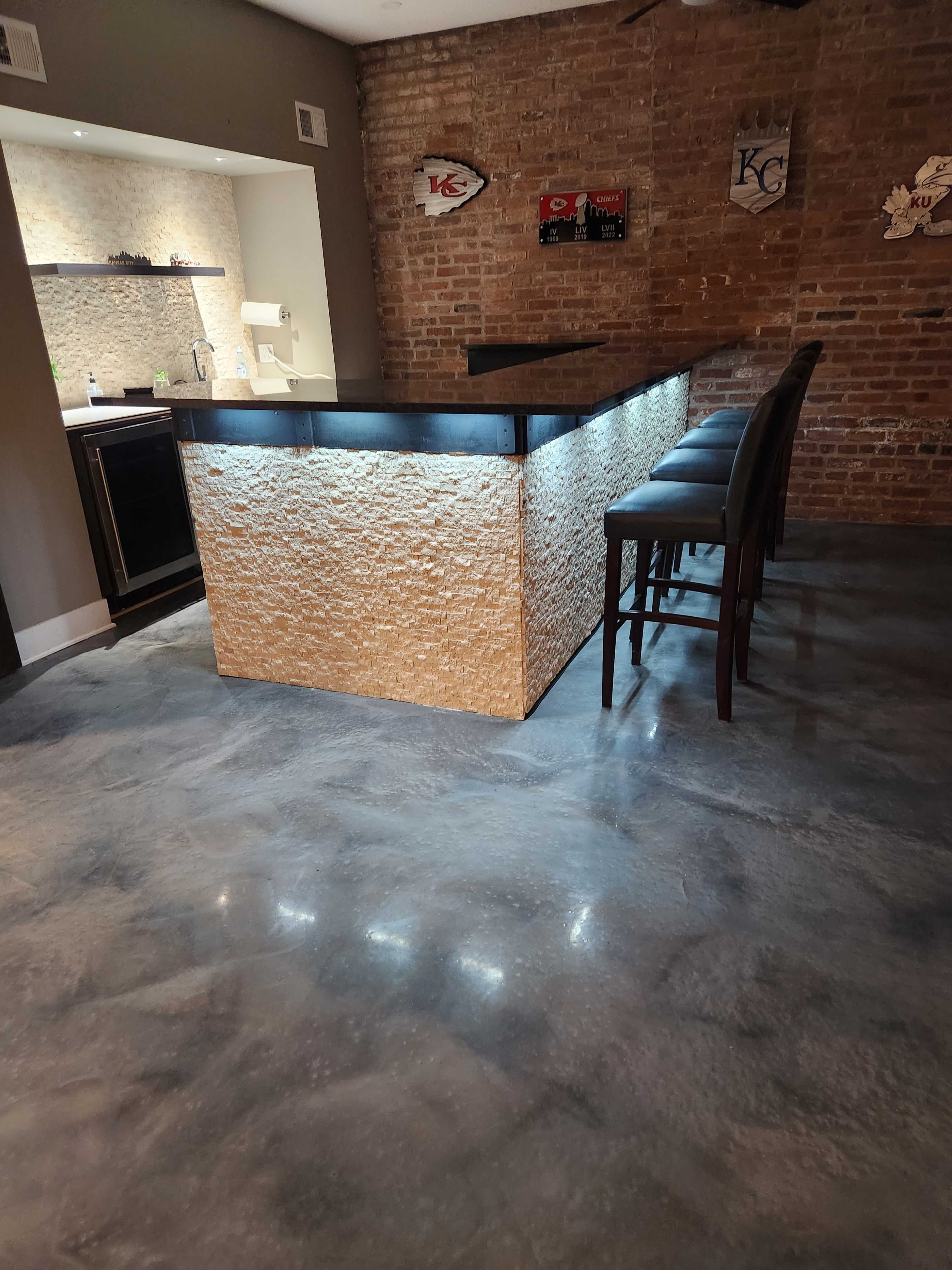 A modern bar area with a stone countertop and backlit base, set against a brick wall and featuring dark bar stools.