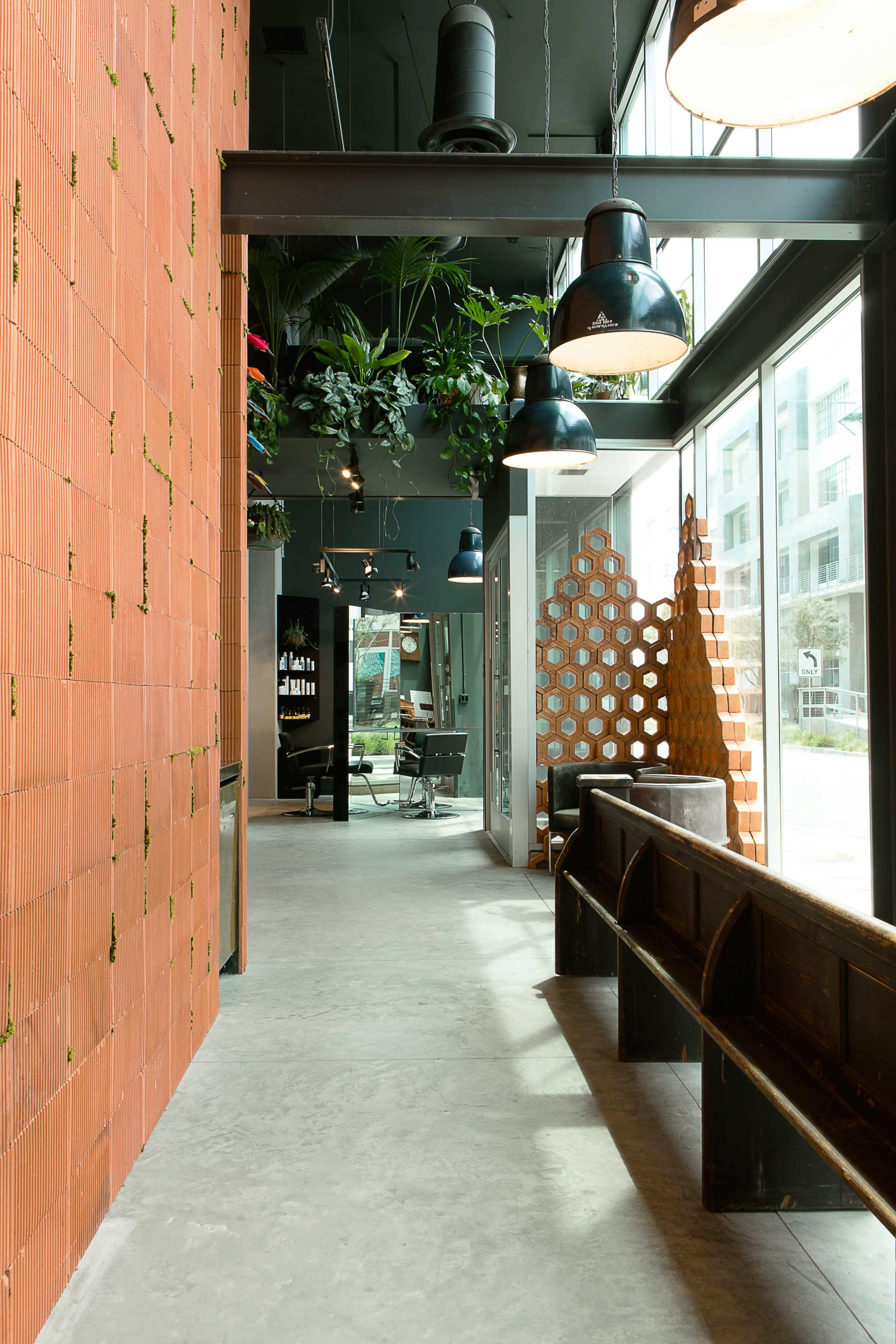 A modern interior hallway featuring a textured orange brick wall, hanging lights, and lush greenery, with a seating area and decorative wooden partition at the far end.