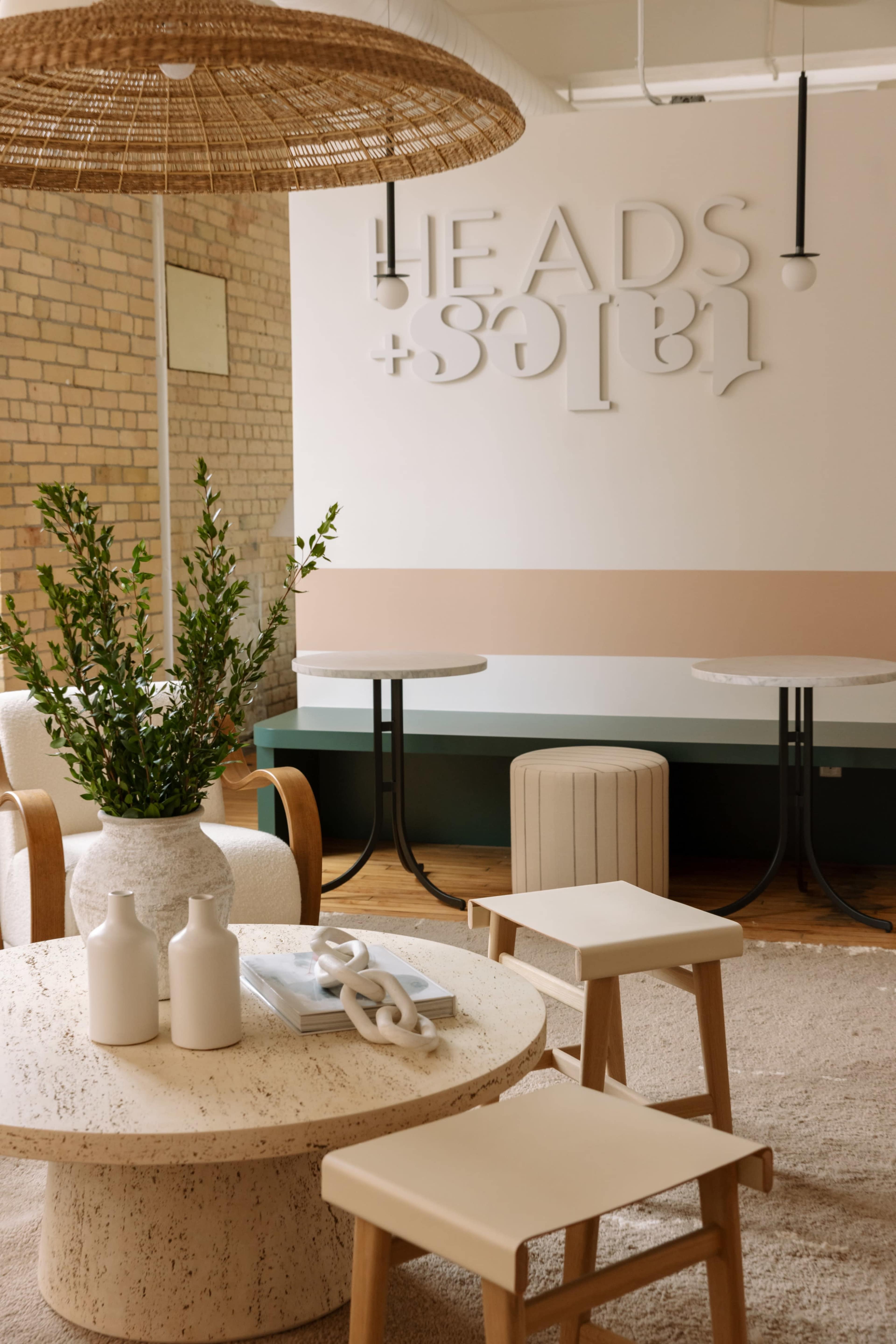 The image features a modern interior space with a round table, minimalist chairs, a potted plant, and a wall displaying the words "HEADS + TAILS" in a stylized design.