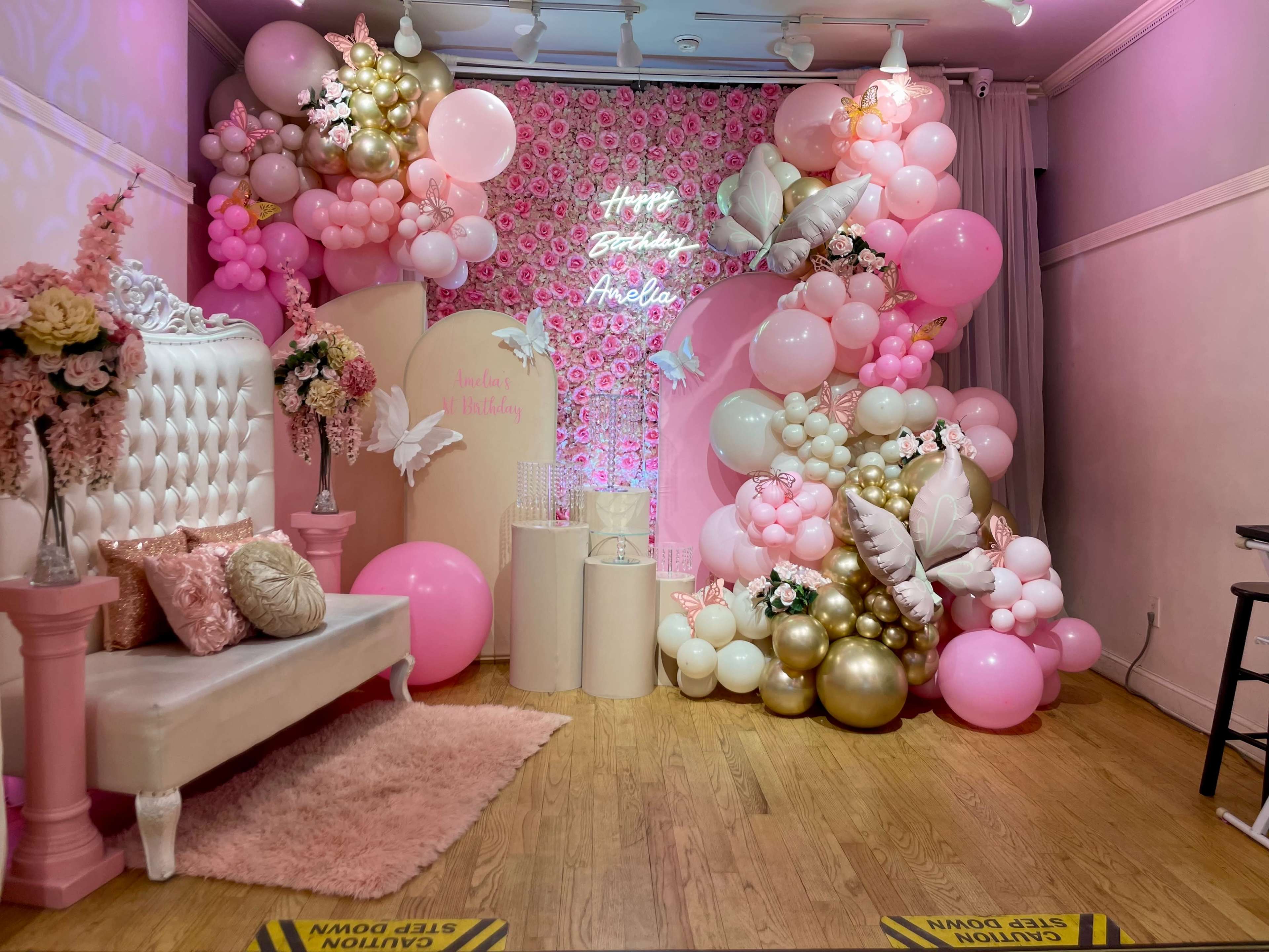 The image features a decorated party area with a white tufted sofa, a backdrop of pink balloons and floral arrangements, and a sign that reads "Happy Birthday," creating a festive atmosphere.