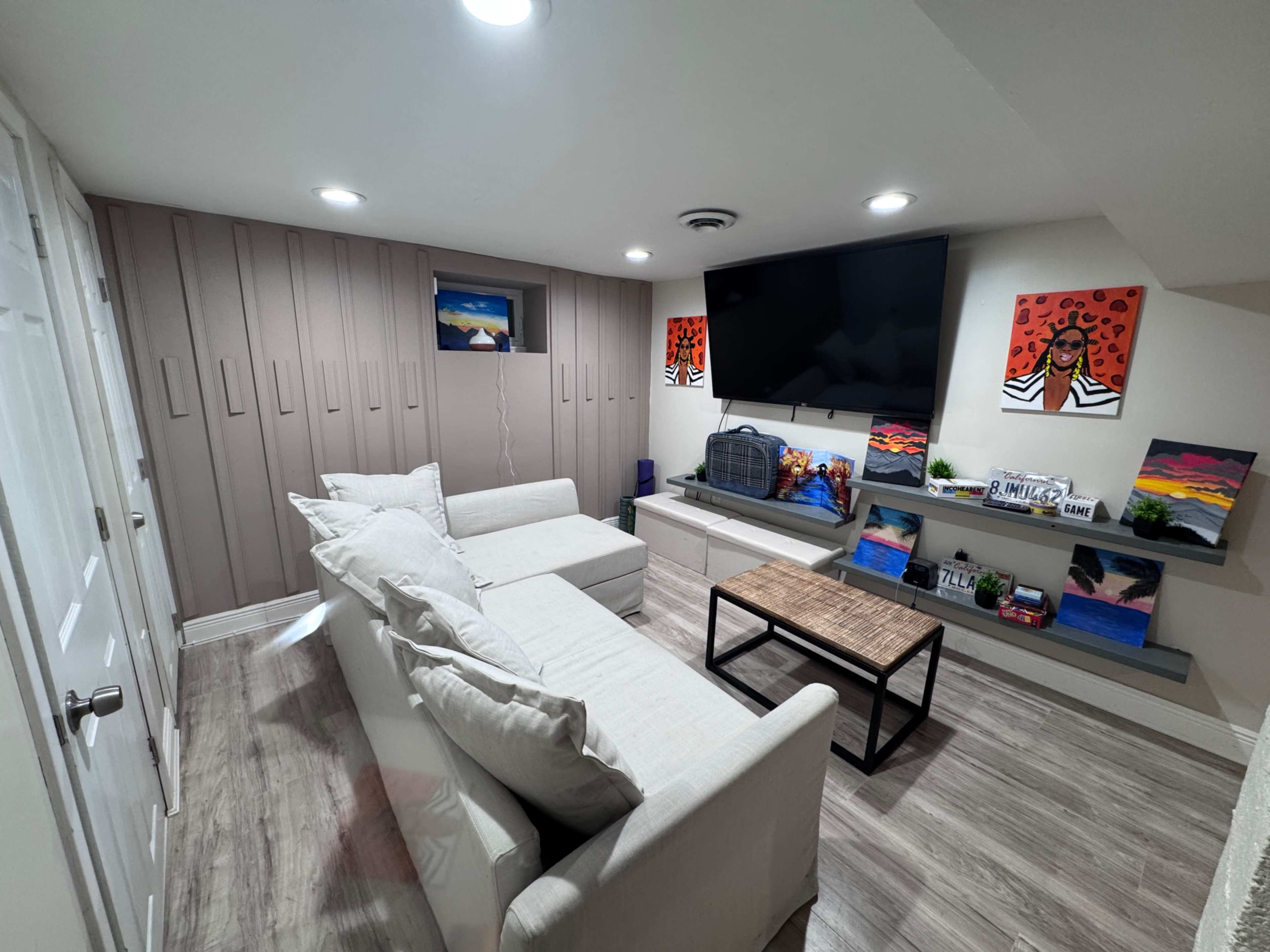 The image shows a modern basement living area with a white sofa, a coffee table, a wall-mounted television, and colorful artwork displayed on shelves.