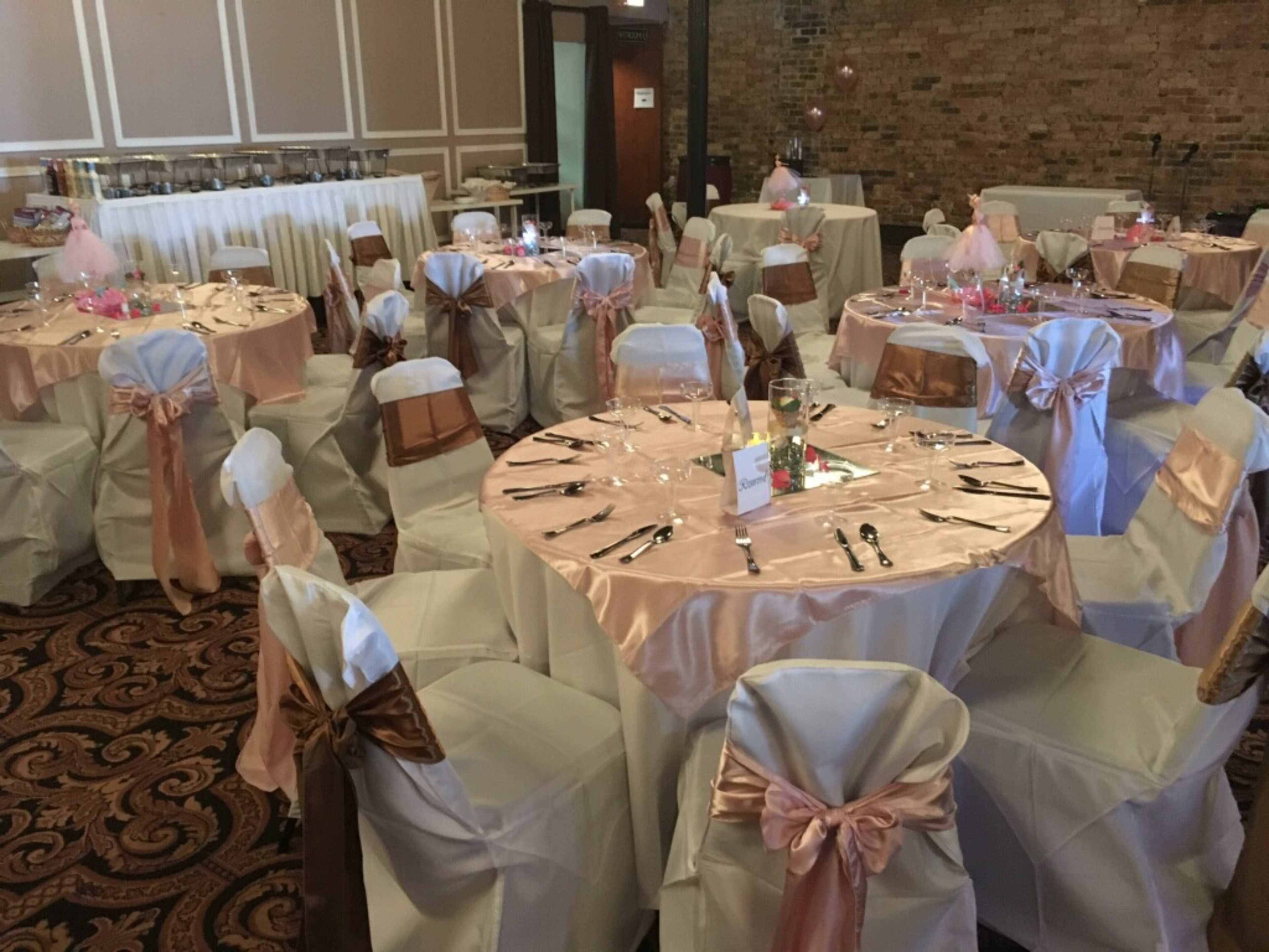 The image shows a banquet hall set up for an event, featuring round tables dressed in pink and gold tablecloths with white chairs adorned in matching sashes.