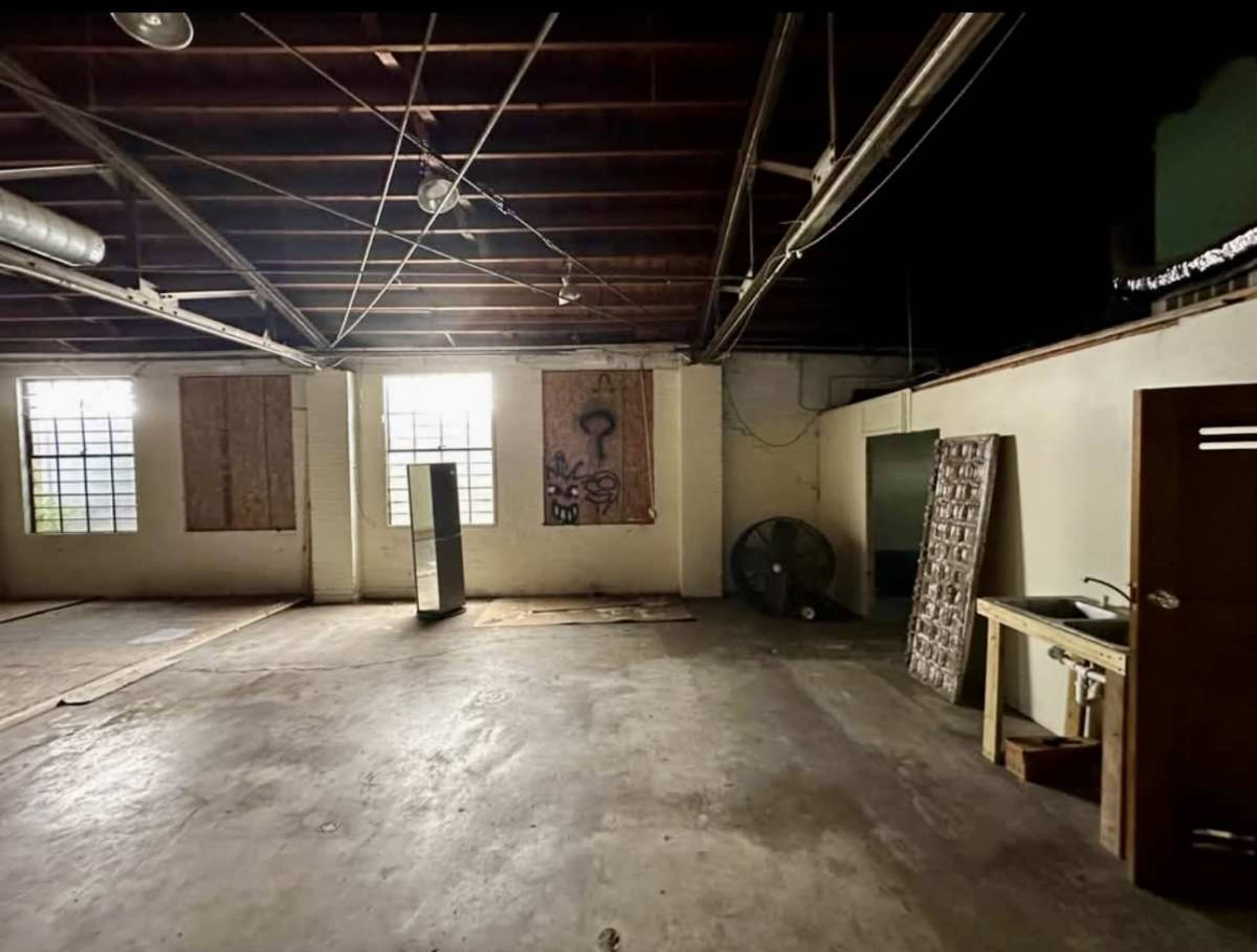 The image shows an empty room with exposed beams, plywood-covered windows, and a large fan in one corner.