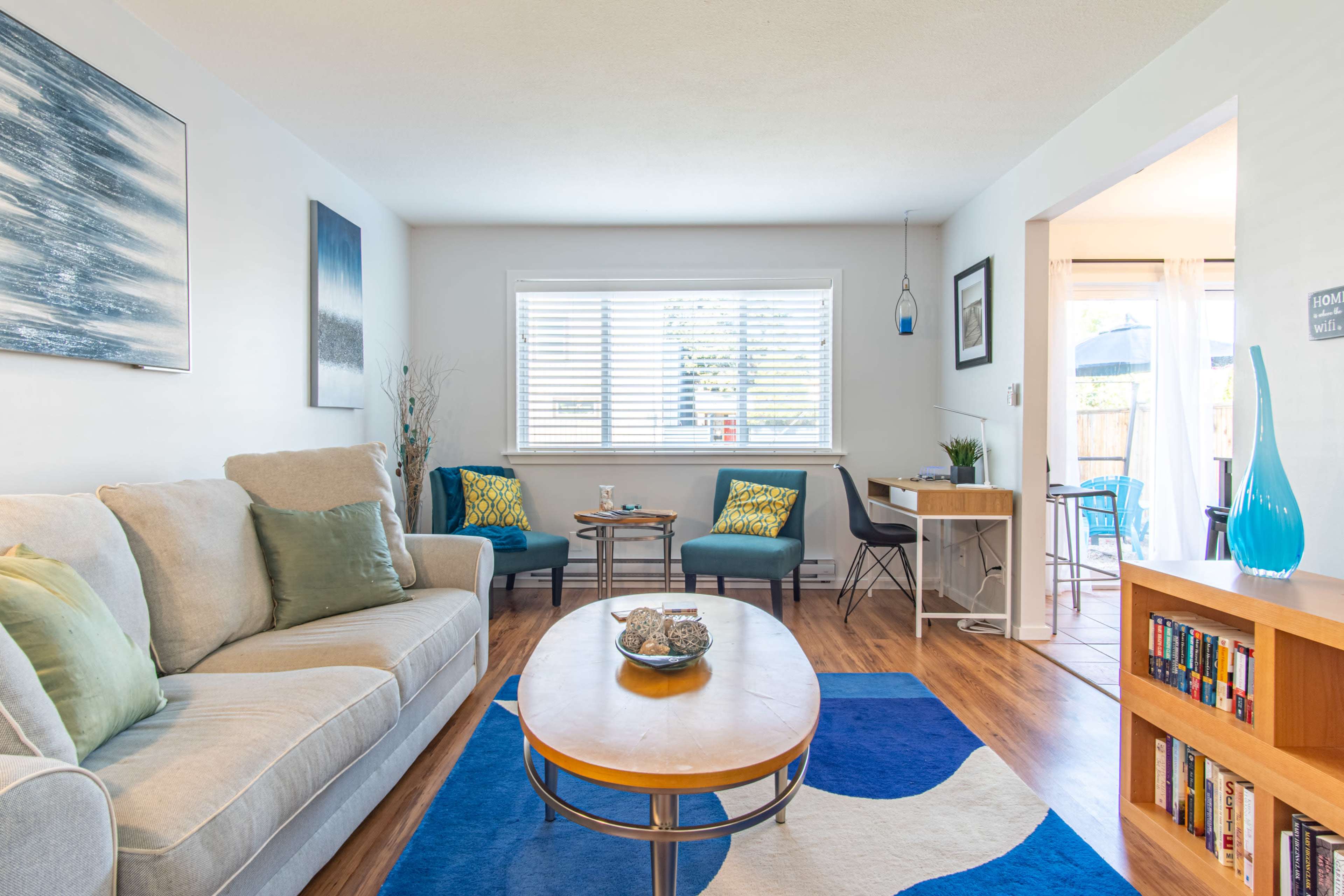 A bright living room with a sofa, a wooden coffee table, and two chairs beside a window, complemented by a small dining area in the background.