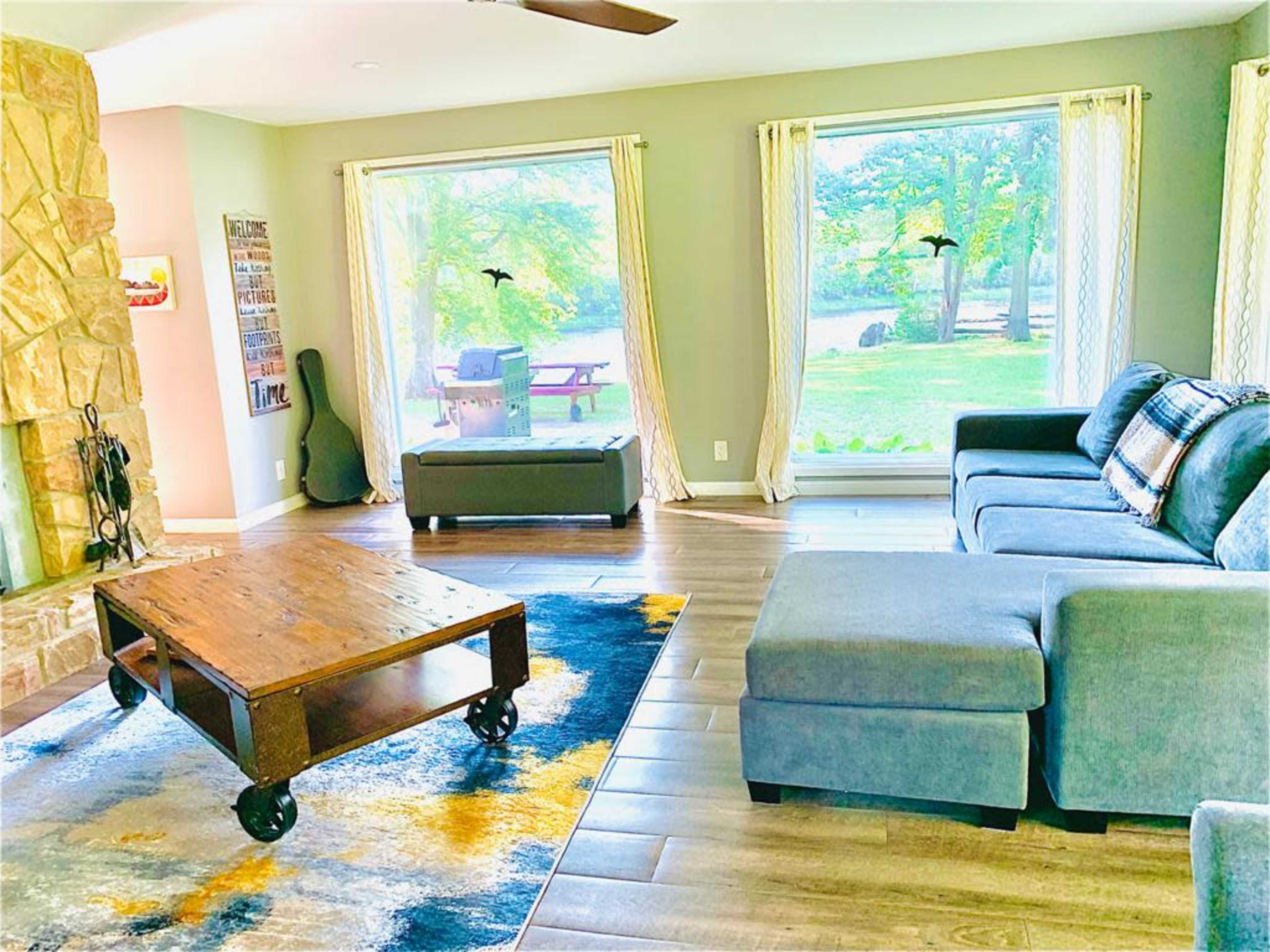 A living room features a stone fireplace, a large gray sectional sofa, a wooden coffee table on a patterned rug, and large windows overlooking a green yard with picnic tables.