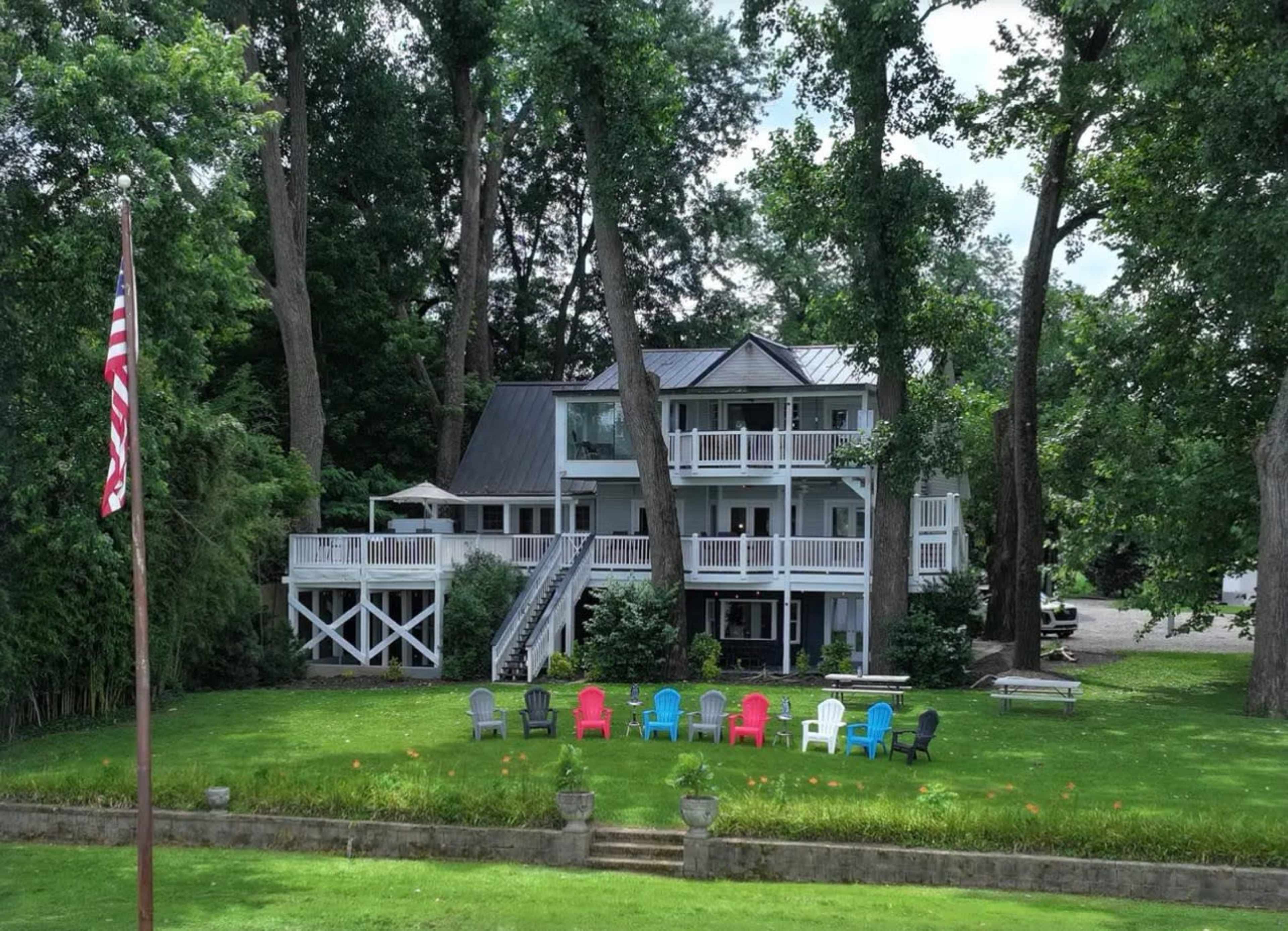 A two-story house with a deck and multiple seating areas is surrounded by tall trees and a green lawn.