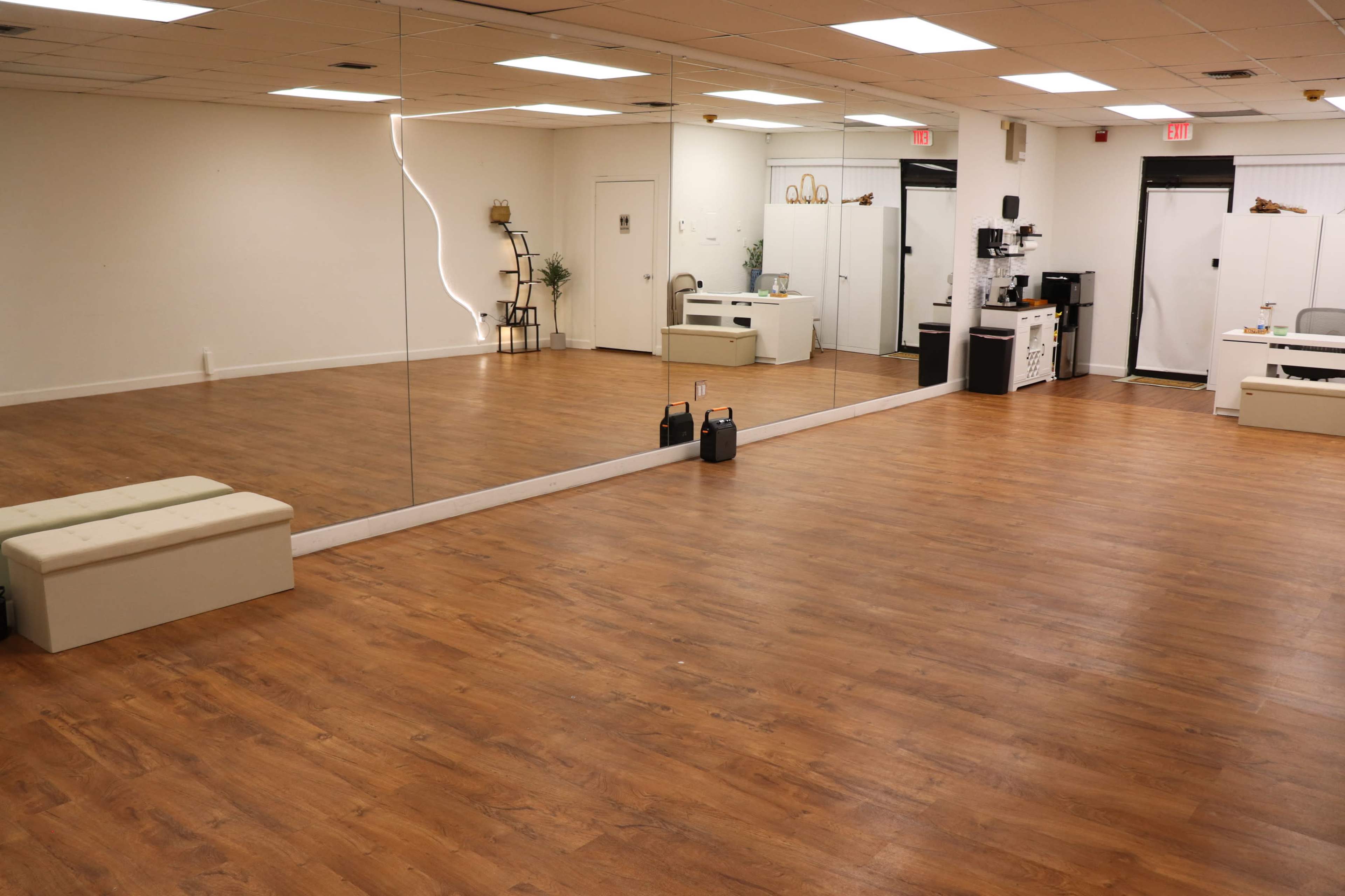 The image shows a spacious, empty room with wooden flooring, a long mirror on one wall, and simple furniture in the opposite corner.