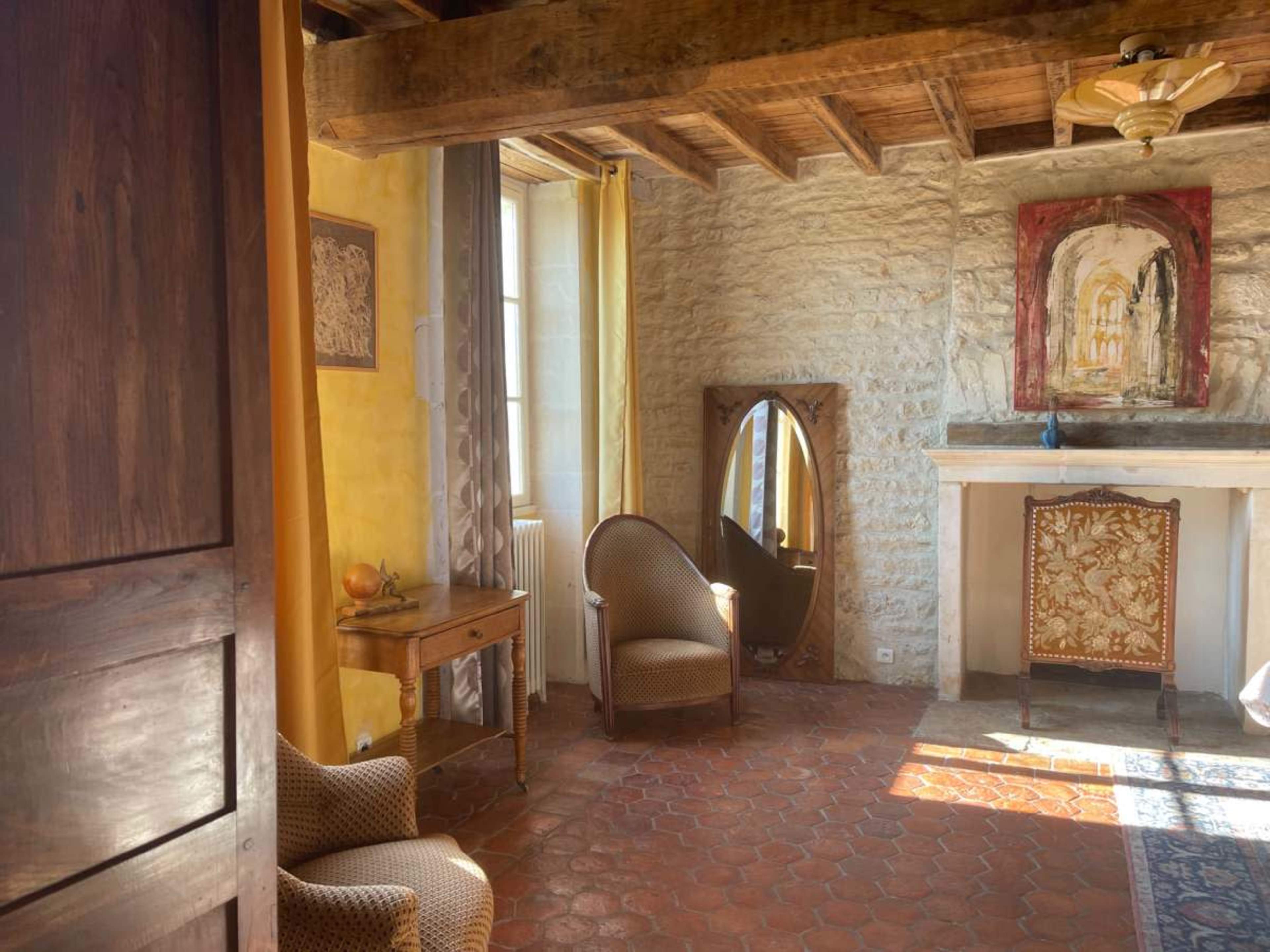 The image depicts a cozy room featuring two chairs, a wooden table, a mirror, a fireplace, and artwork on the stone wall.
