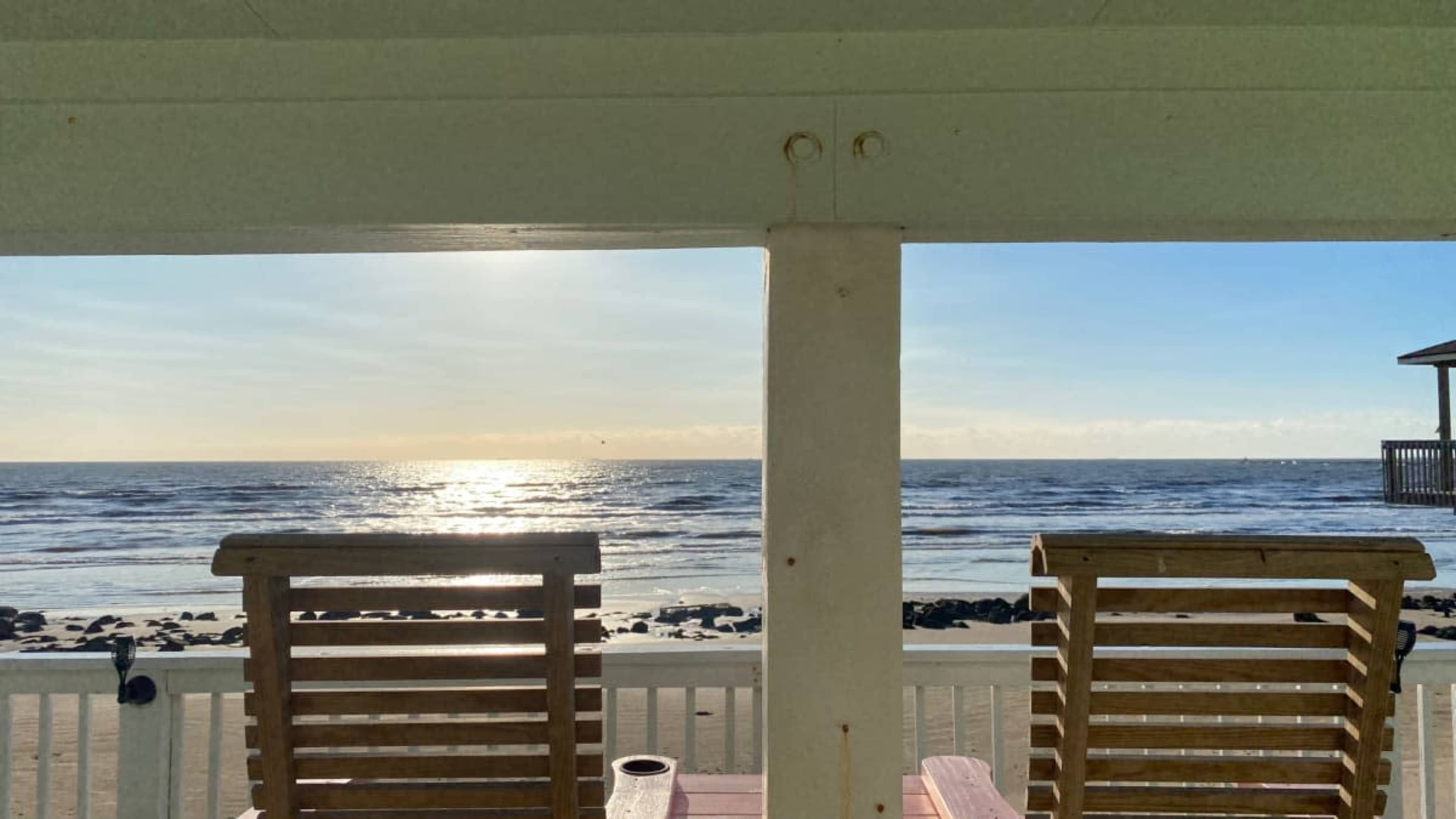 The image shows two wooden chairs facing a view of the ocean and a sunset over the water, framed by a white porch railing.