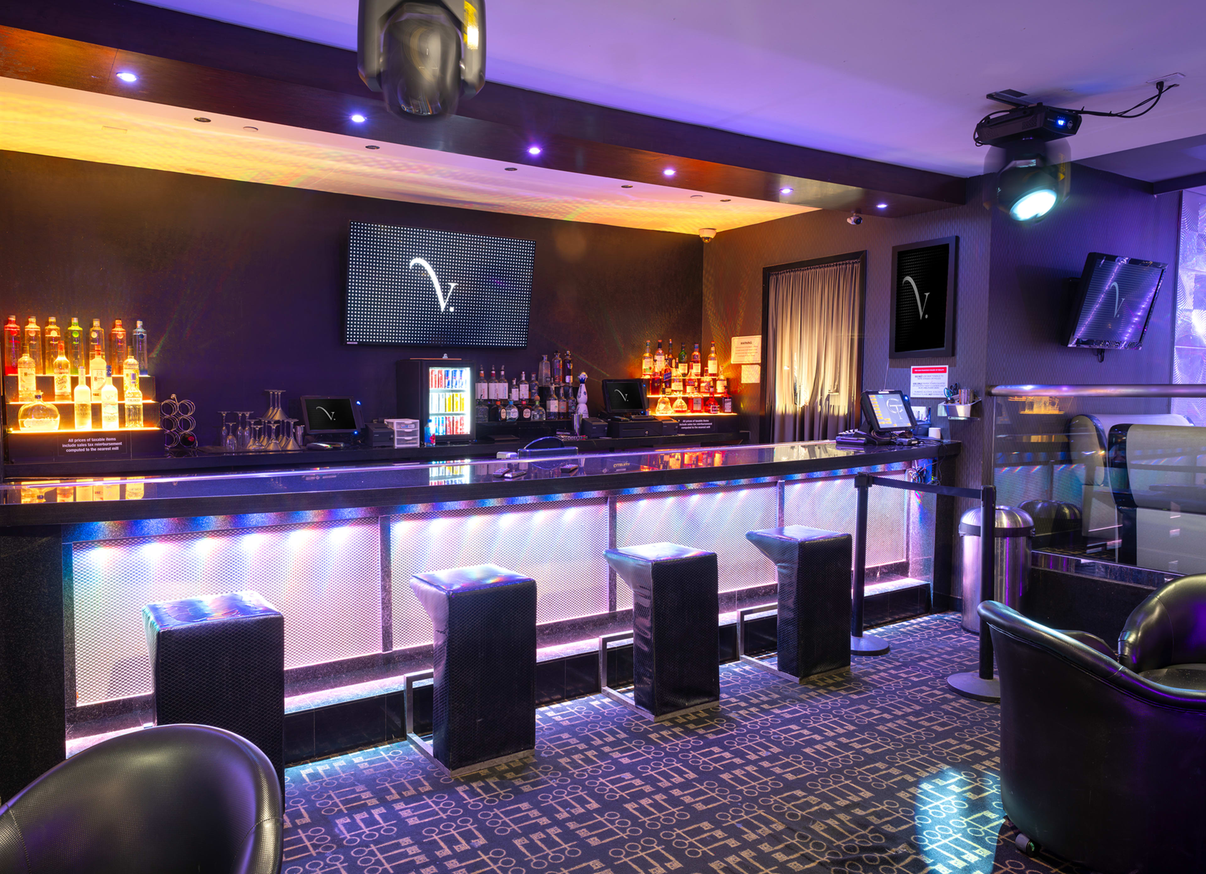 The image shows a modern bar area with illuminated shelves of liquor, a sleek counter, and several black stools arranged in front.