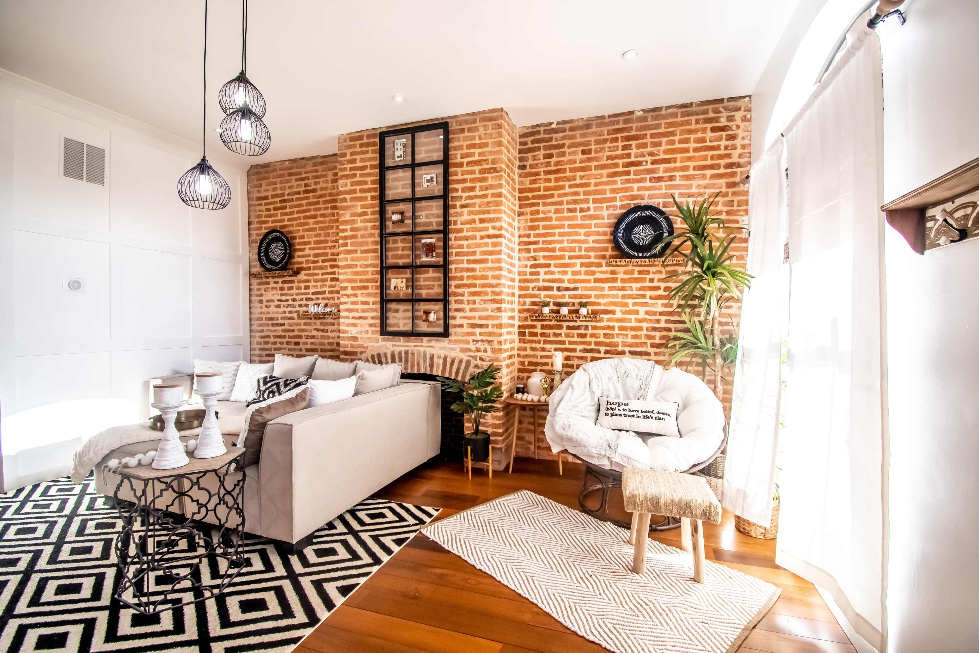 The scene features a modern living room with a brick accent wall, a light-colored sectional sofa, a patterned rug, and decorative plants.