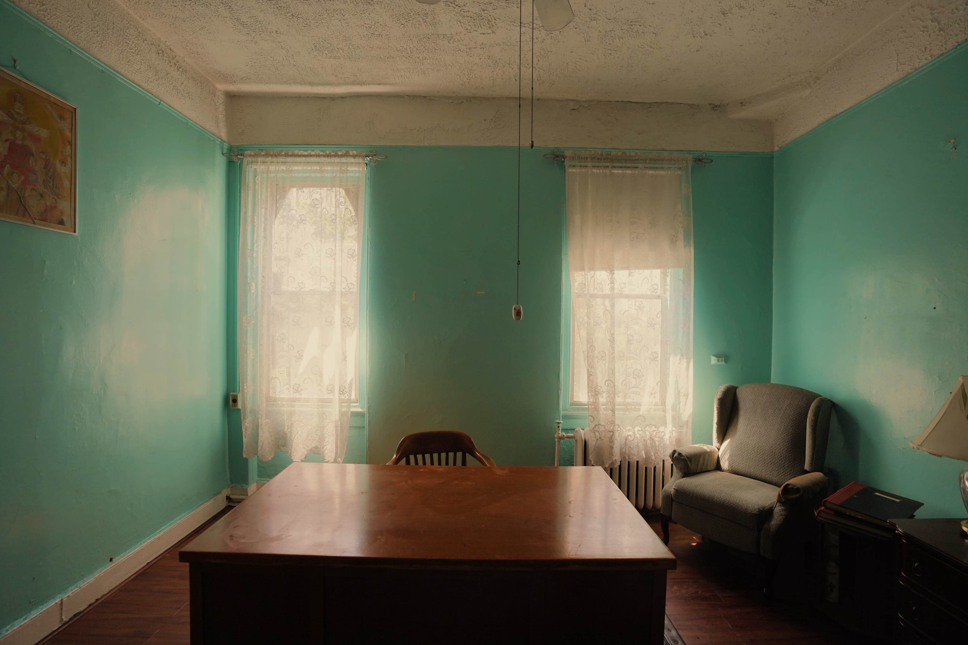 A room with turquoise walls, a large wooden table in the center, two windows with white lace curtains, and a chair beside a gray armchair.