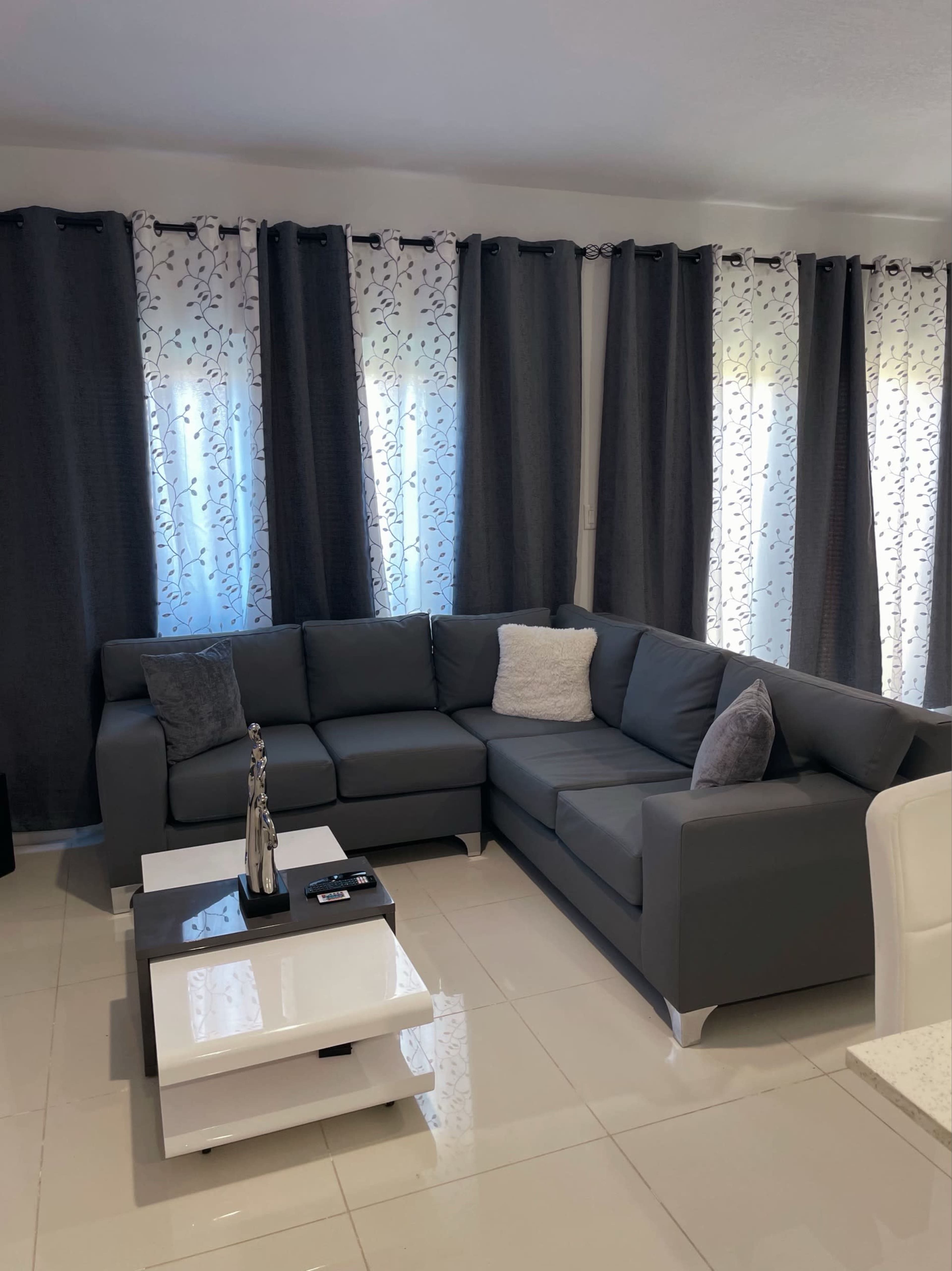 A modern living room with a gray sectional sofa, a white coffee table, and patterned curtains allowing natural light to filter in.