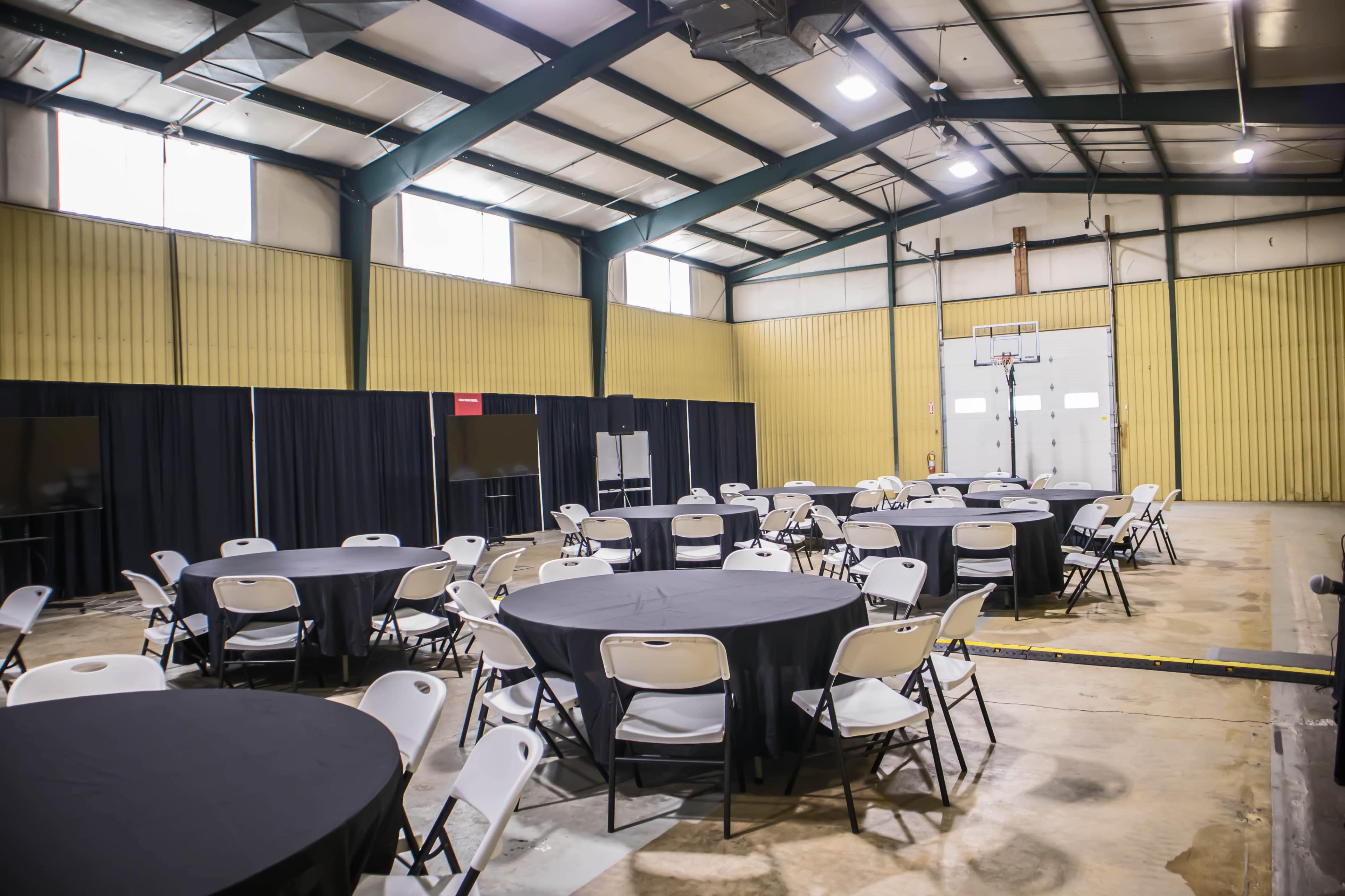 A spacious indoor hall features circular tables surrounded by chairs, with black drapes and a basketball hoop at the far end.